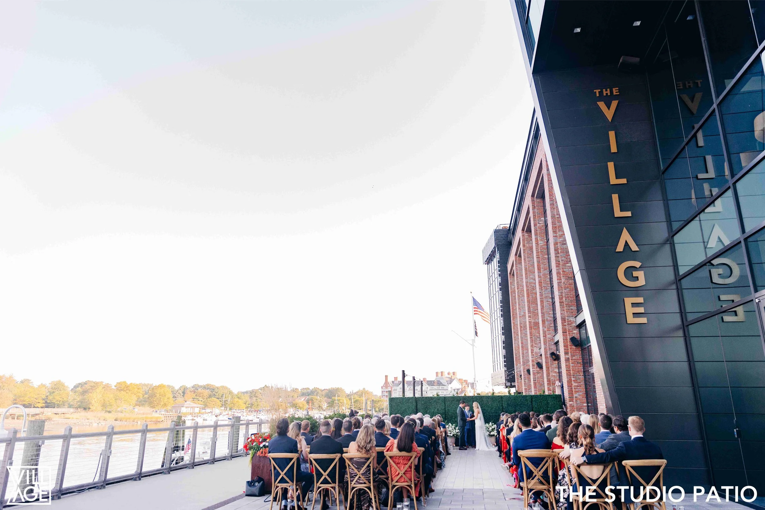 Outdoor wedding ceremony on a patio by the water, with guests seated facing an officiant and bride and groom, near a modern building with large glass windows and a sign that reads 'The Village.'