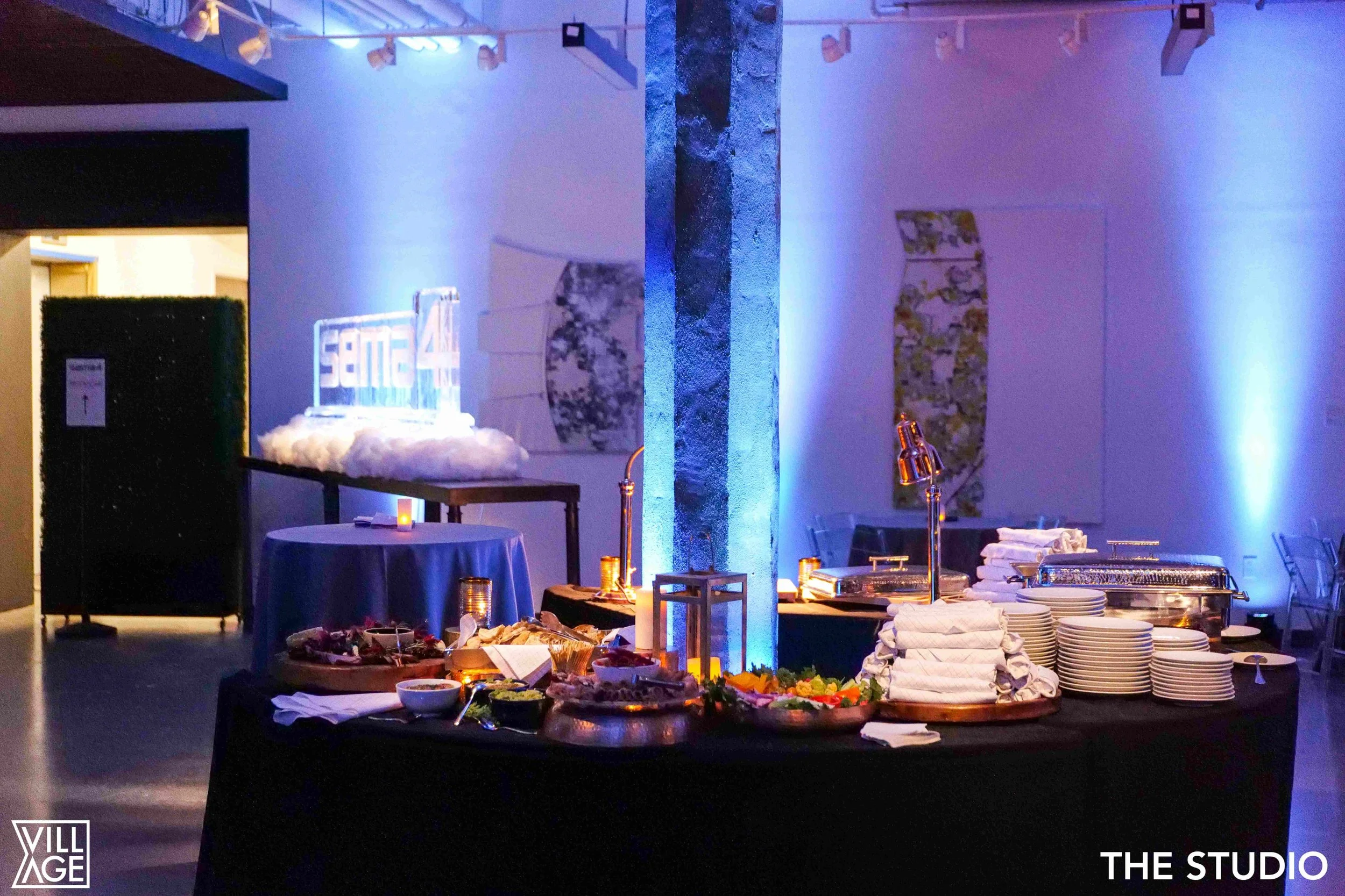 Buffet table with salads and chafing dishes in a dimly lit room with blue lighting, likely at a party or event. There is a central pillar with blue LED lighting and a neon sign in the background.