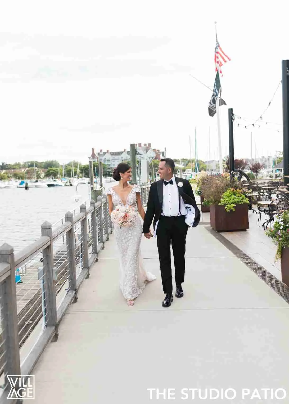Bride and groom walking hand in hand along a waterfront patio, smiling at each other, with sailboats and buildings in the background.