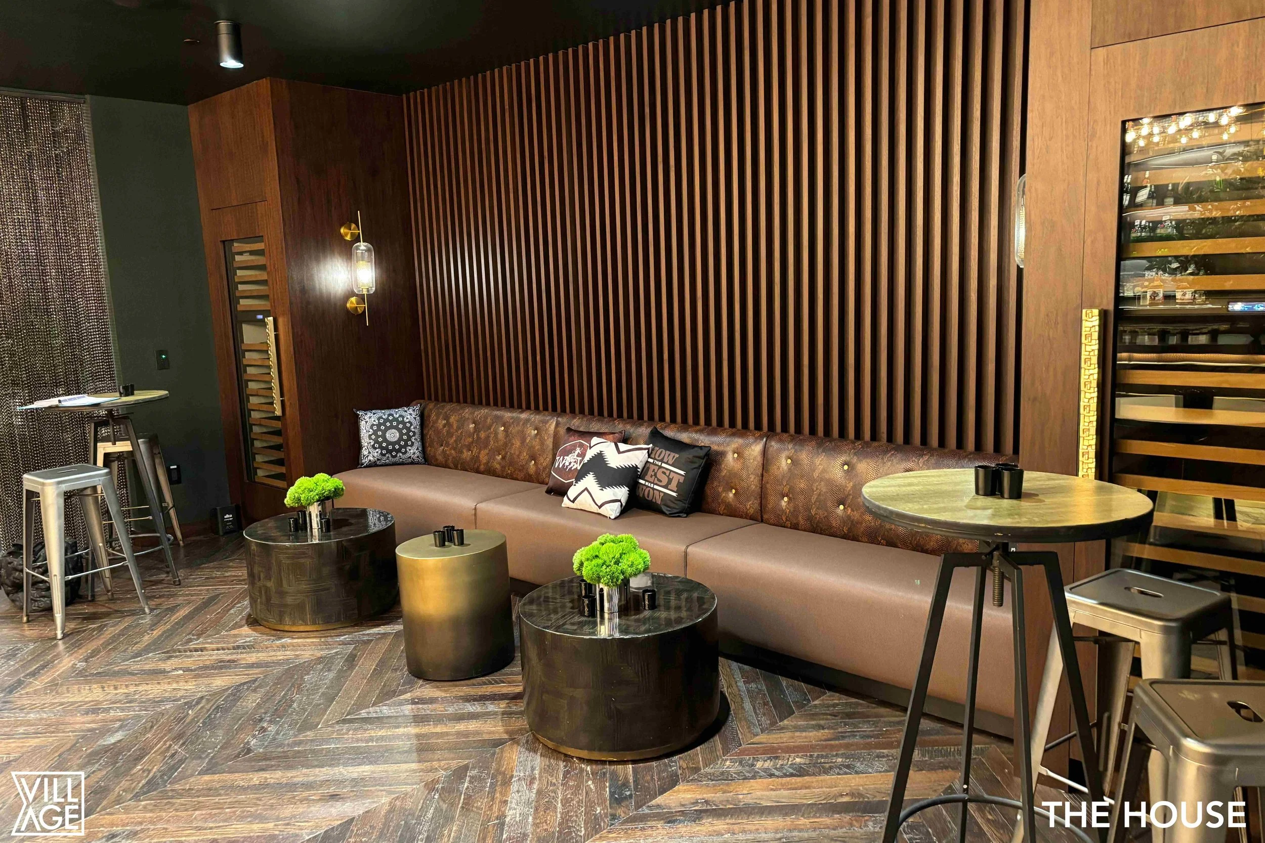 Modern lounge area with long beige sofa, black marble coffee tables, decorative pillows, wooden wall paneling, small round tables with green plants, and metal bar stools.