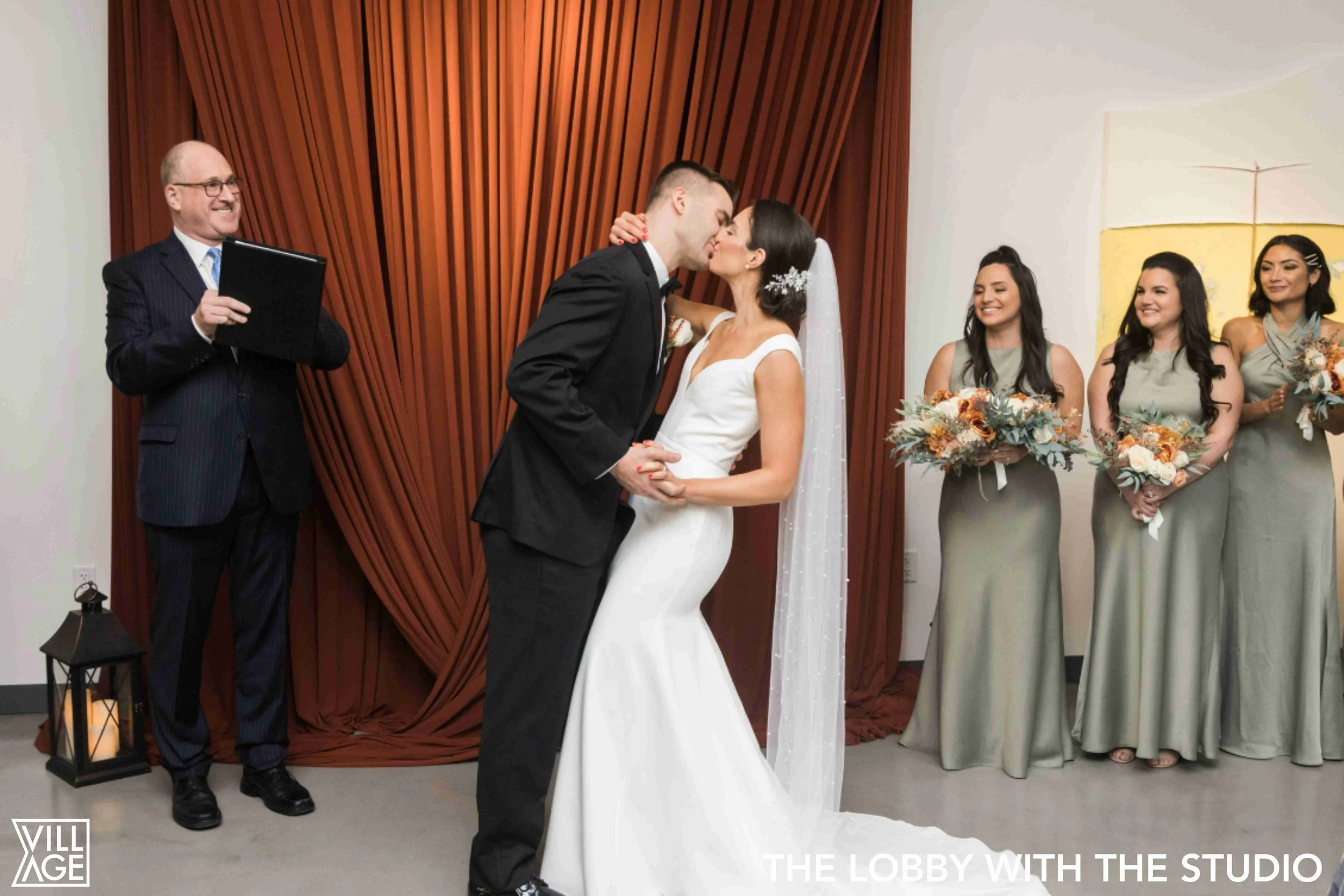A bride and groom kiss during their wedding ceremony, with bridesmaids holding bouquets in the background and an officiant watching smiling.