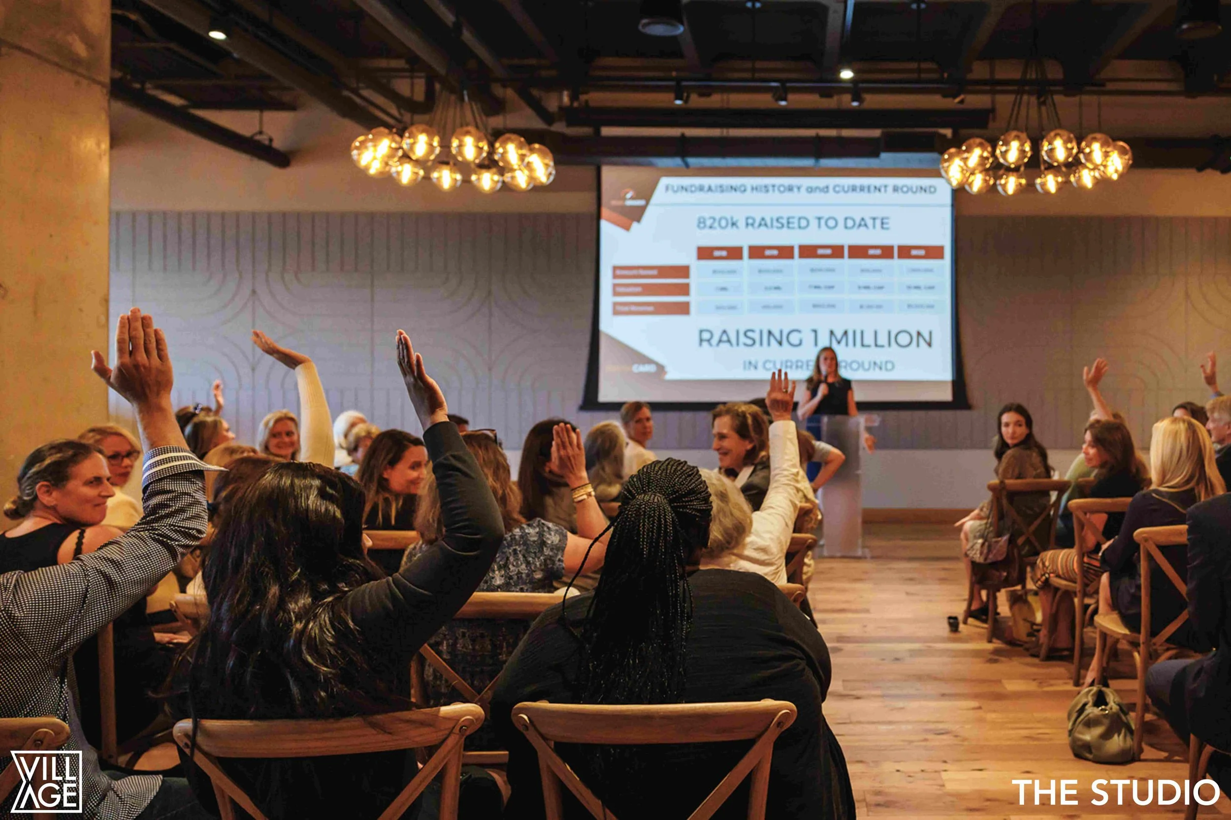 A group of people seated in a conference room, raising their hands to ask questions or participate in a discussion, with a presenter at the front and a large screen displaying fundraising progress in the background.