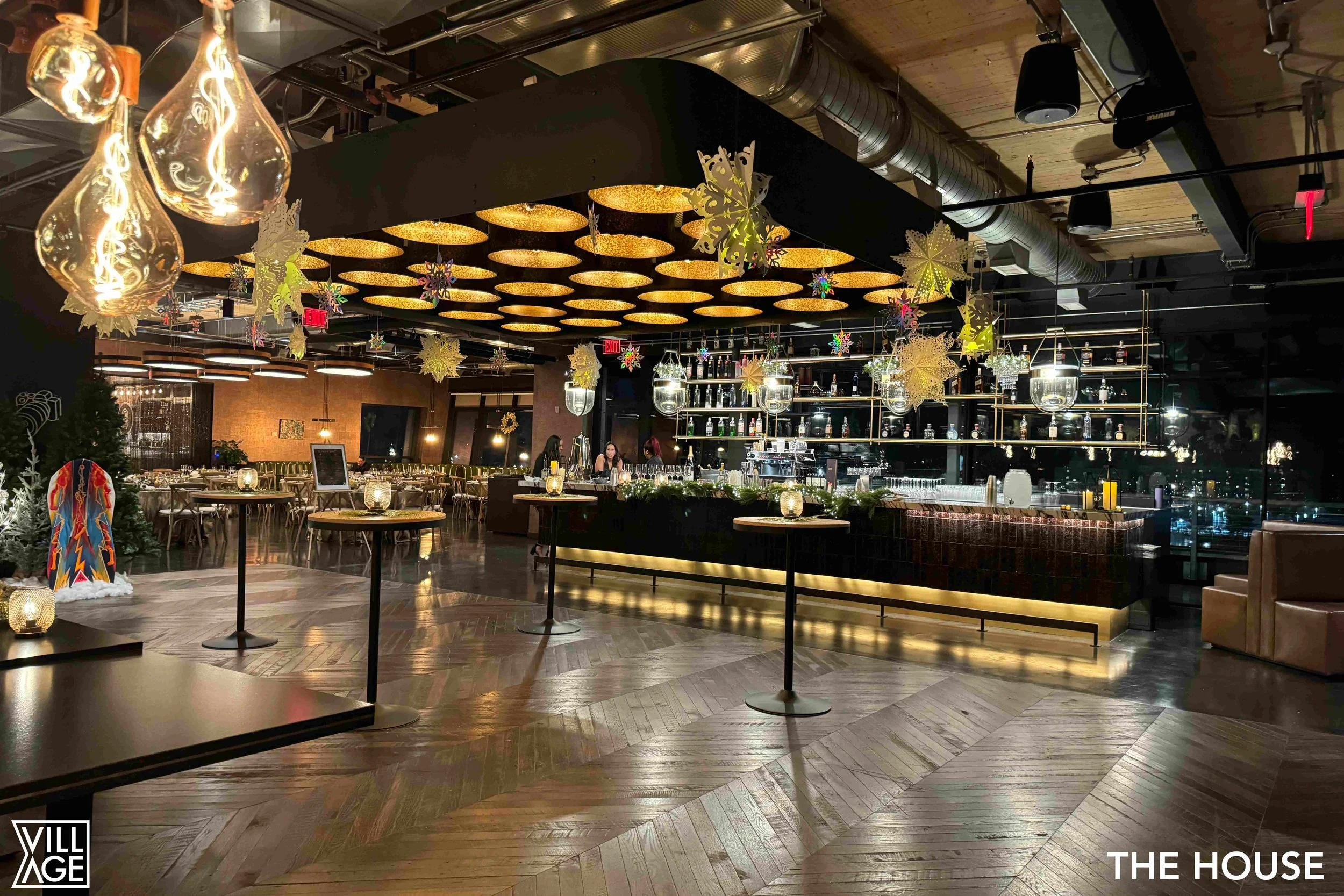 Interior of a dimly lit bar and restaurant decorated for the holidays with hanging snowflakes and ornaments, a lit bar counter, and tables with candles, in a modern industrial style.