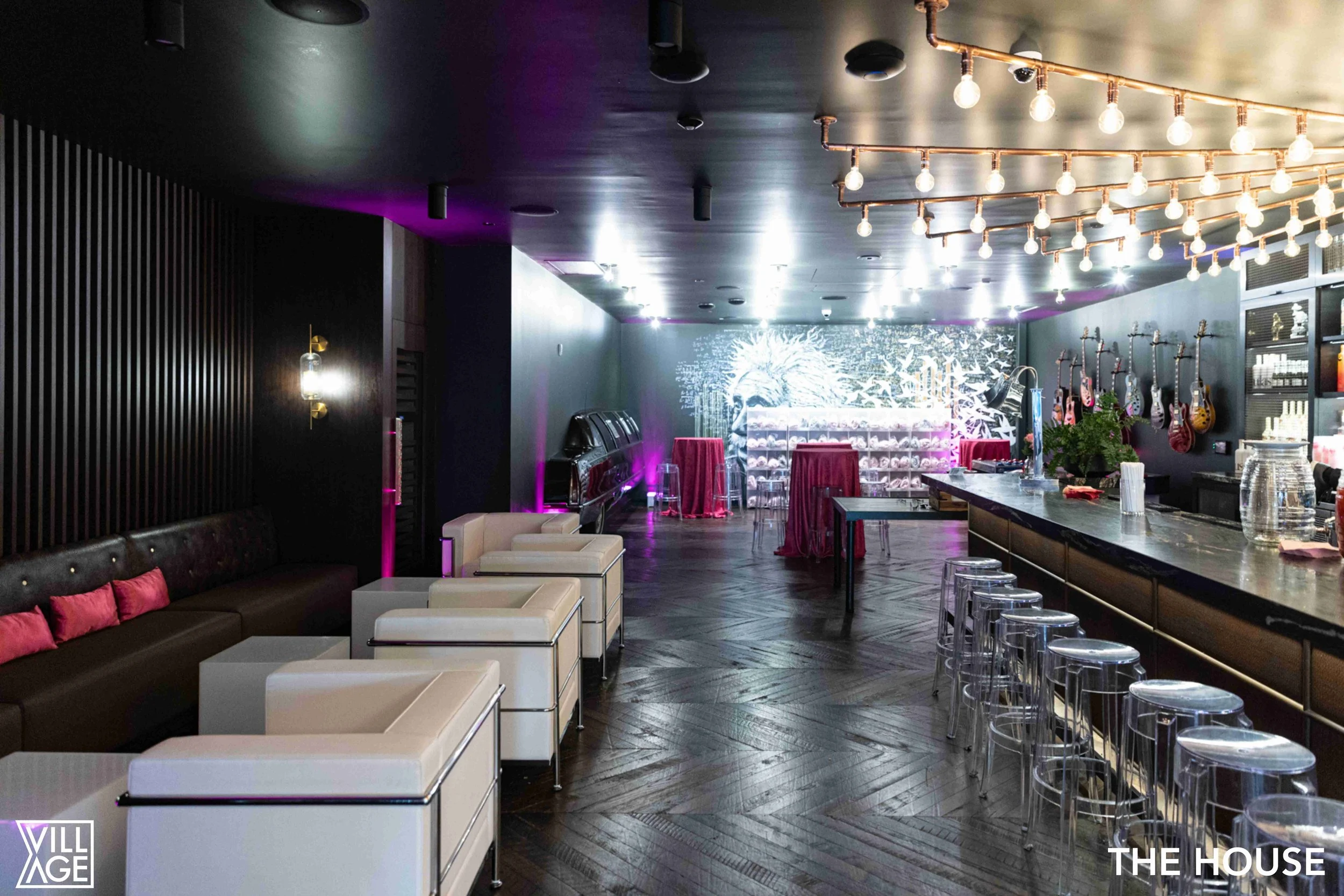 Interior of a bar or lounge with modern decor, black and white furniture, pink accents, hanging light fixtures, guitars on the wall, and a large mural in the background.