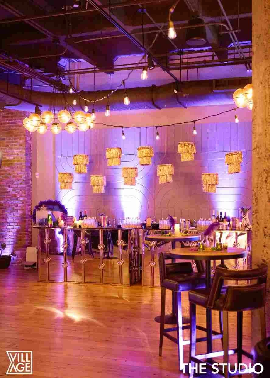 Indoor event space with purple lighting, string lights, hanging lamps, a bar with glasses and bottles, and high chairs around small tables.