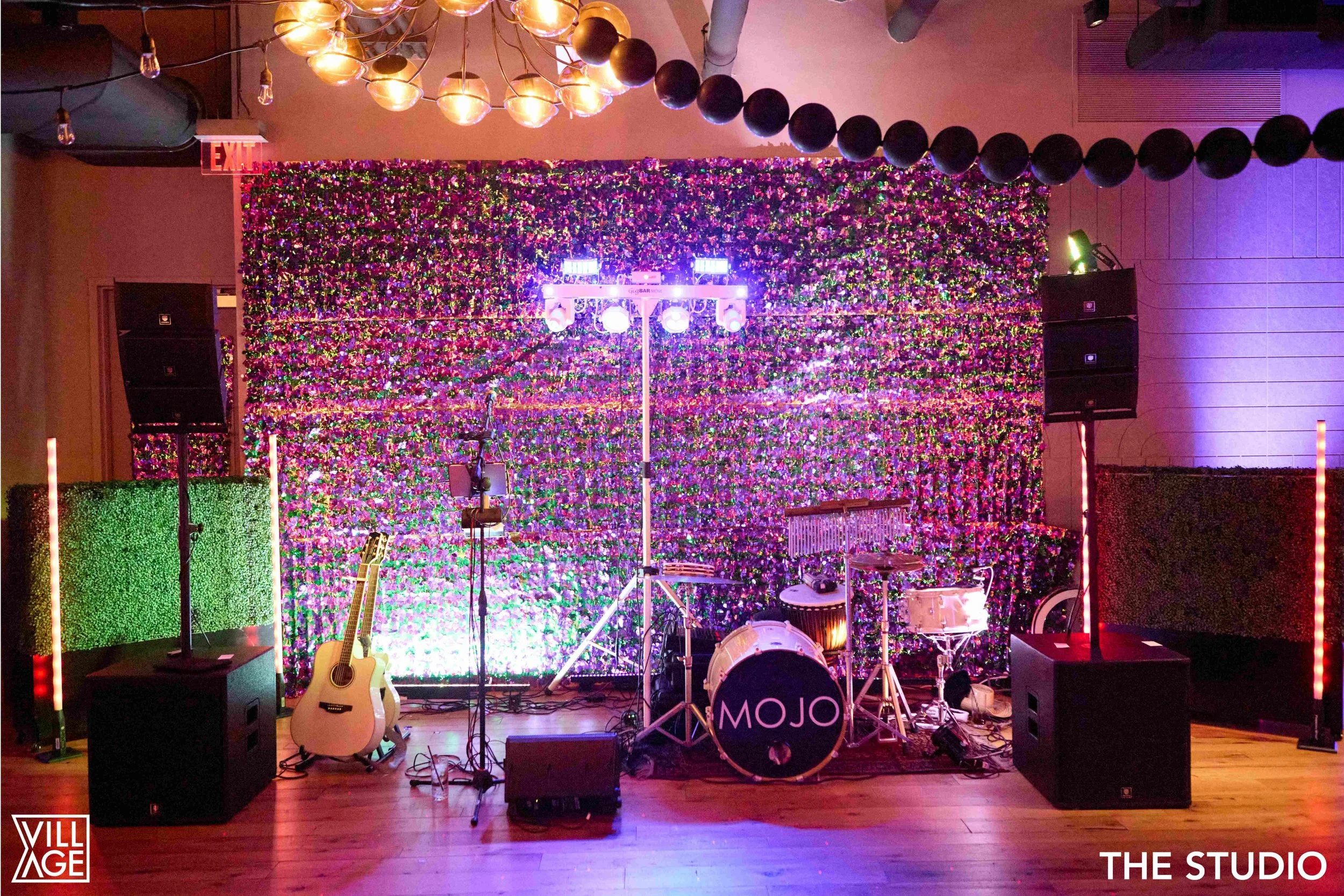 A stage setup with musical instruments including a drum set with the word 'MOJO' on the bass drum, an acoustic guitar, and a microphone stand with music sheets, all in front of a decorated floral backdrop. The stage is lit with colorful lights and ha