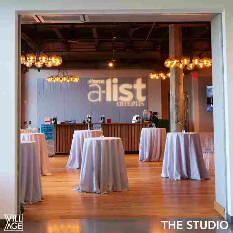 Interior of a event space with high ceilings, hanging globe lights, round tables with pink tablecloths, and a bar area in the background with a wall projection reading 'the a-list (nights)'.