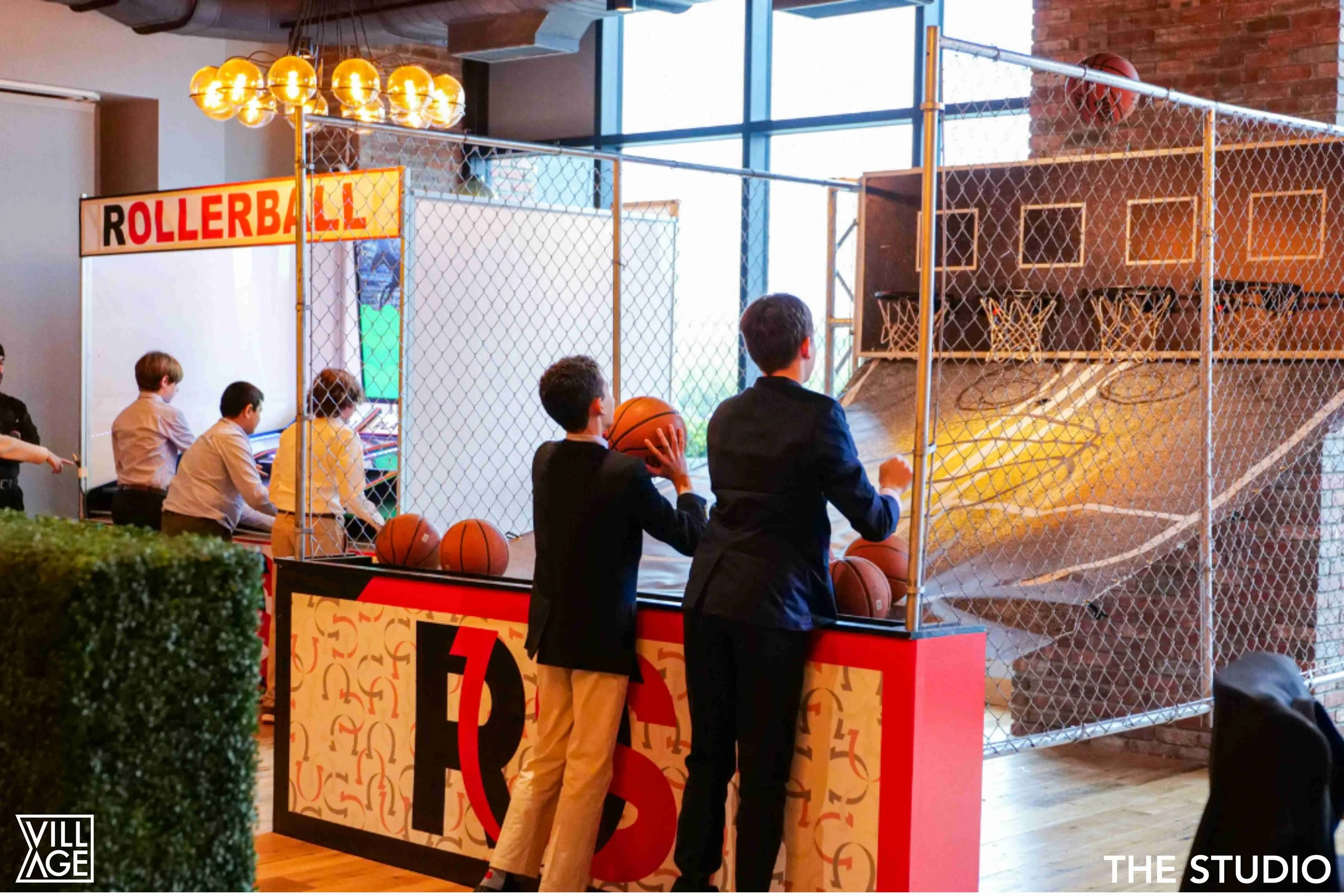 Indoor arcade with children playing basketball at a roller skating rink enclosed in a chain-link fence, with a volcano-shaped slide in the background.