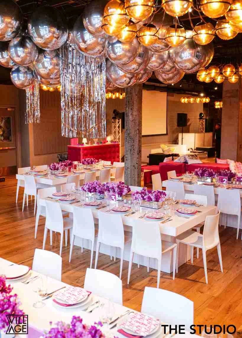 Decorated event space with tables set for a celebration, pink floral centerpieces, metallic balloons, hanging silver streamers, and a projection screen in the background.