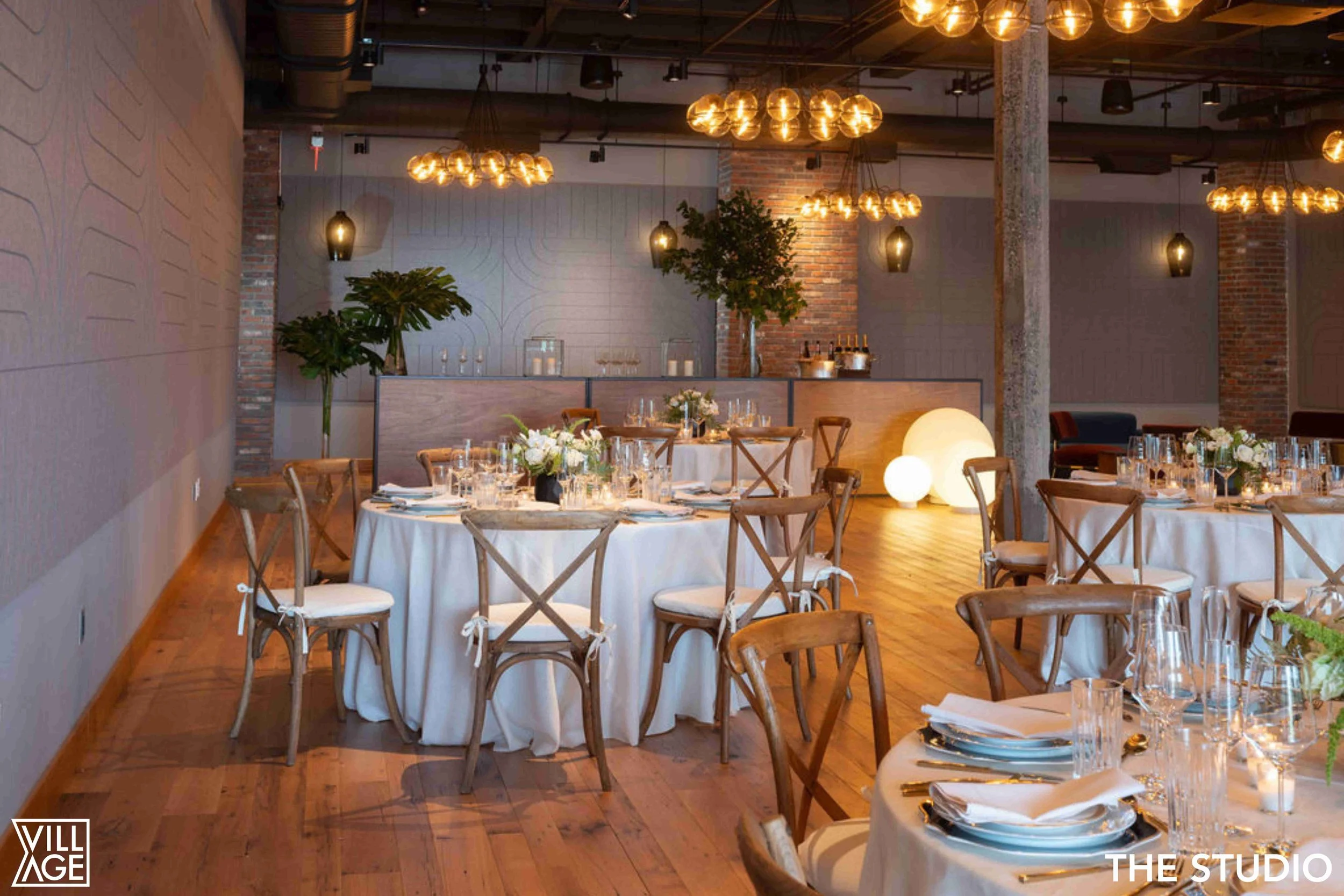 Elegant indoor event space with round tables covered in white tablecloths, set with glassware, plates, and silverware. Wooden chairs surround the tables. Warm lighting from modern pendant and bulb chandeliers, with decorative glowing orbs on the floo