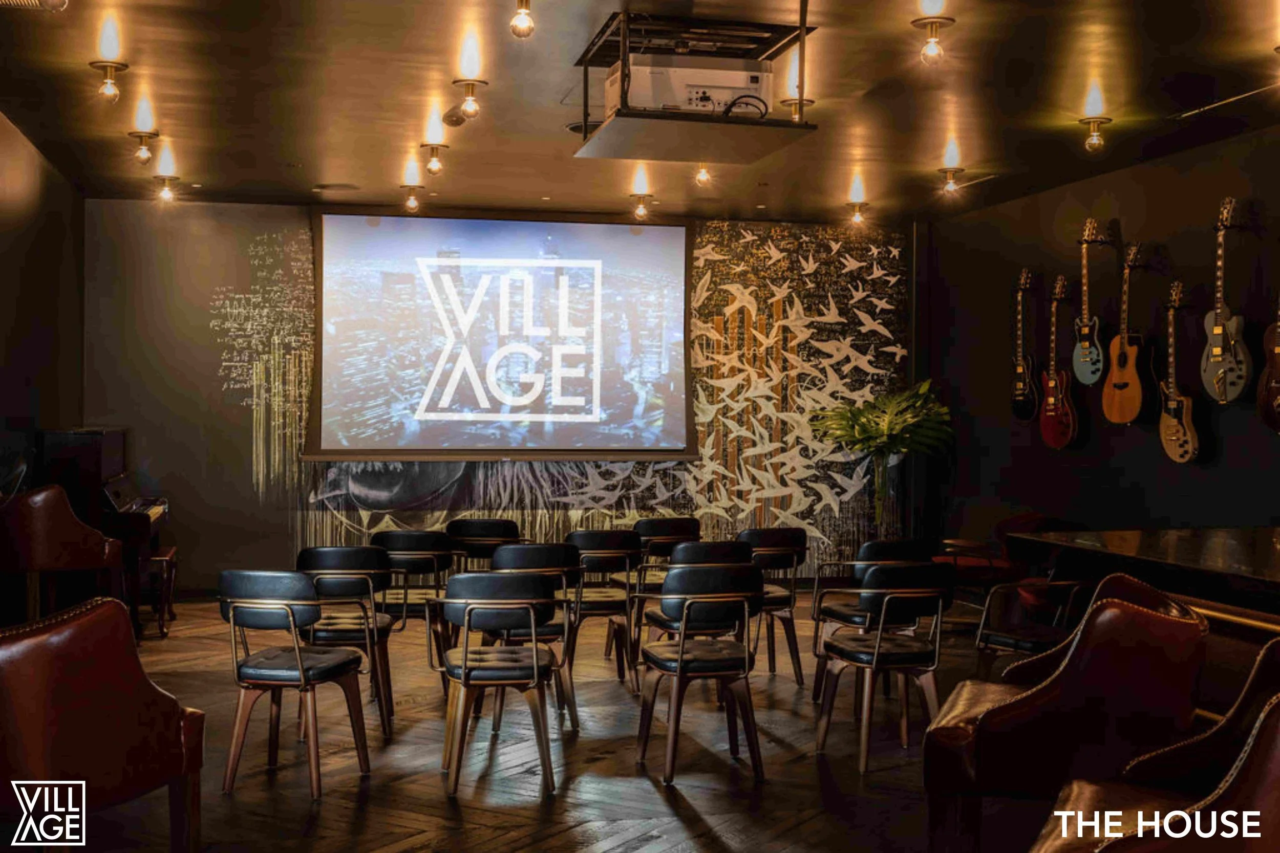 Inside a dimly lit room with chairs set up facing a large screen displaying the word 'VILLAGE' with a cityscape background. The room features musical instruments on the wall, a potted plant, and a decorative wall of white birds in flight. The ceiling