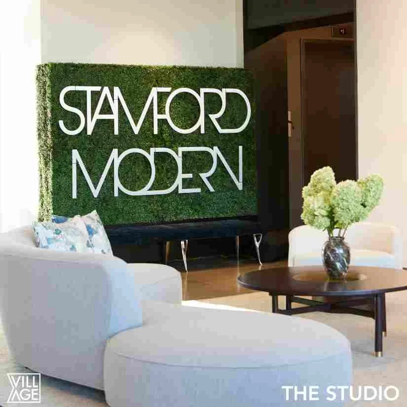 Interior studio space with a sign that reads 'STAMFORD MODERN' on a green artificial grass wall, a white curved sofa with a decorative pillow, a black coffee table with a vase of white hydrangeas, and a white chair nearby.