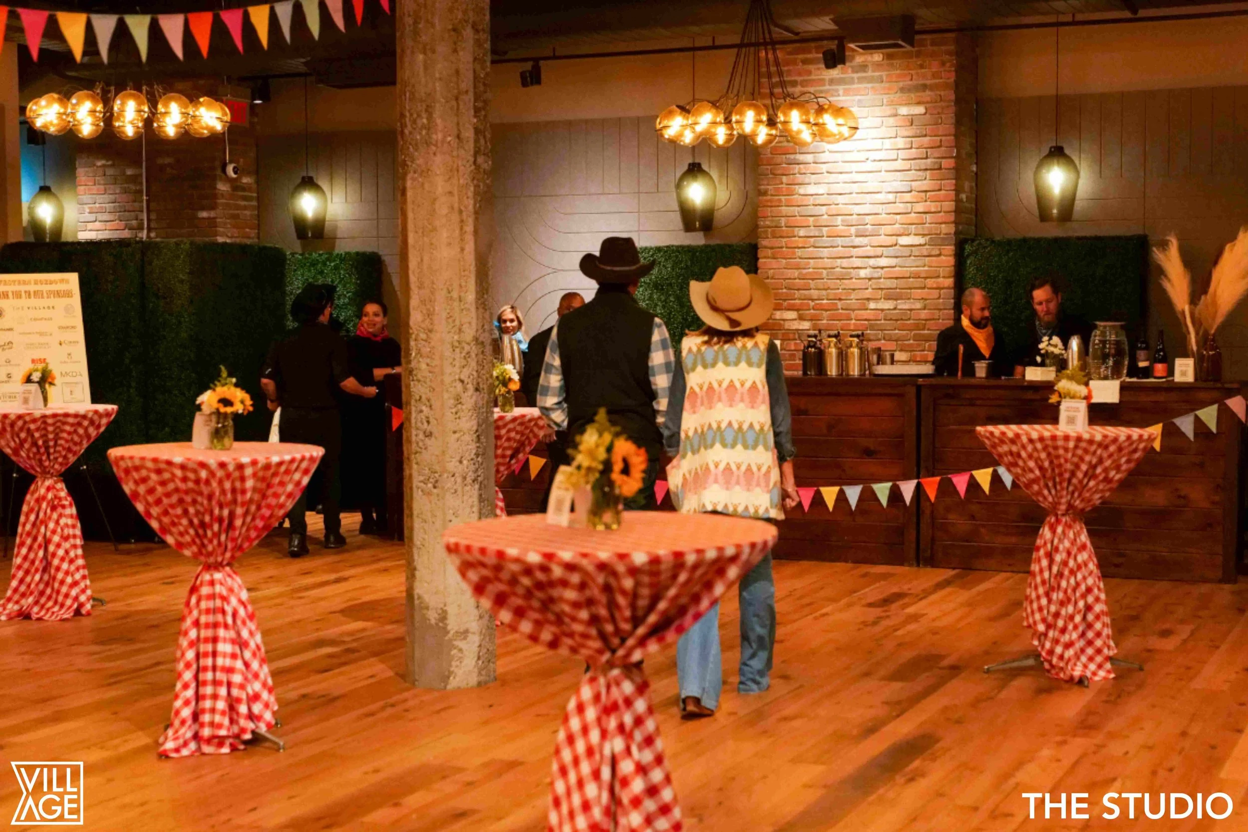 Indoor event space with high tables covered in red and white checkered tablecloths, decorative flowers, string banners, and a bar area in the background, with people dressed in cowboy hats and Western attire.