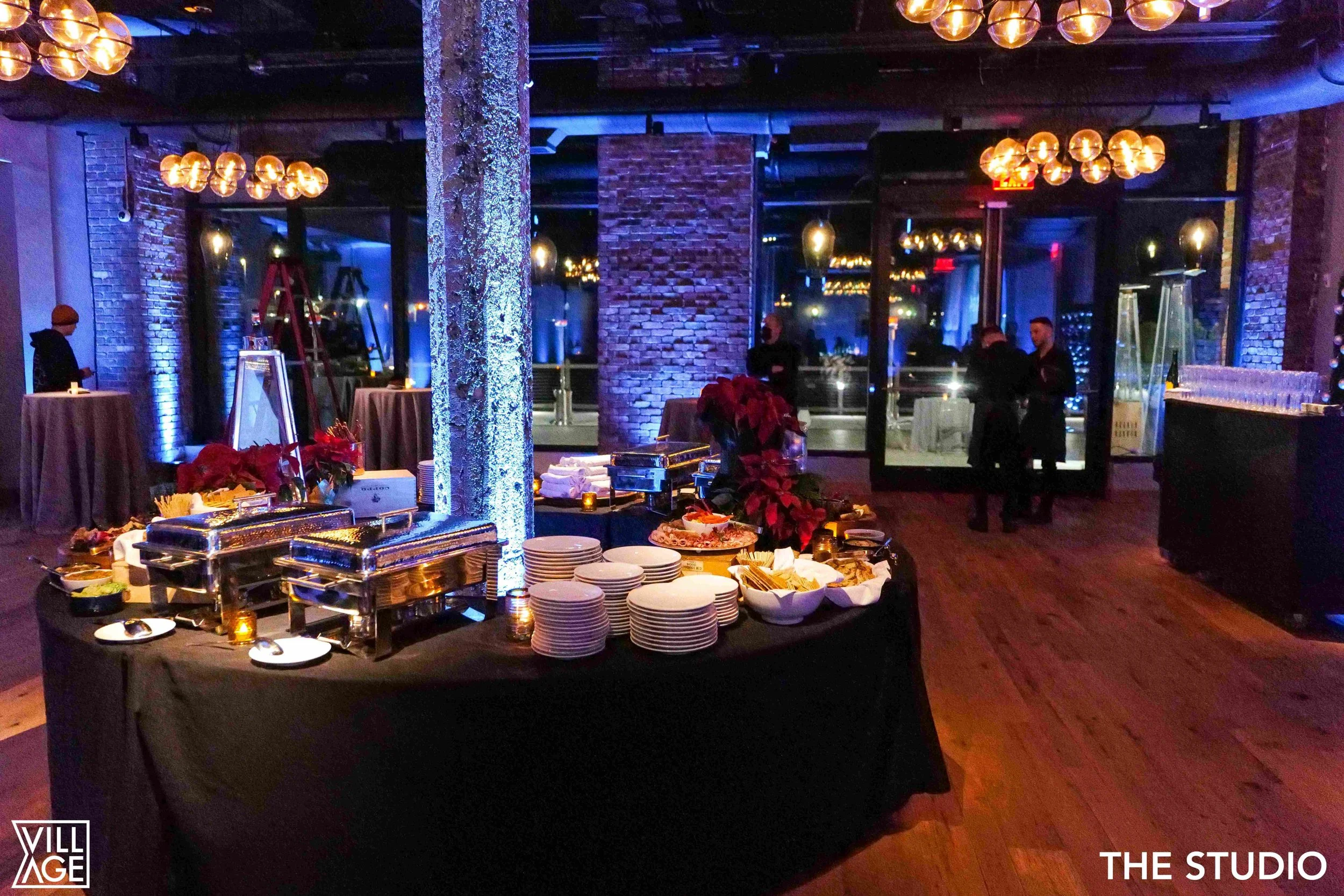 Indoor event space with a buffet table in the foreground, decorated with poinsettias, plates, and chafing dishes. The room features exposed brick walls, large windows, and modern lighting fixtures, with a few people standing and chatting in the backg
