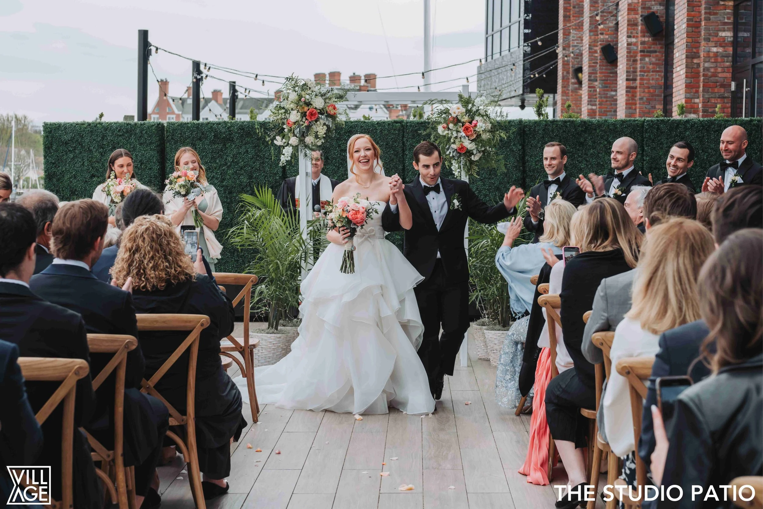 Bride and groom walking down an aisle after wedding ceremony in outdoor patio, surrounded by guests taking photos and clapping, with floral arch and greenery backdrop.
