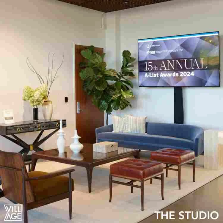 A modern living room with a blue sofa, wooden coffee table, two leather ottomans, a wooden armchair, plants, and a wall-mounted TV displaying the 15th Annual A-List Awards 2024.