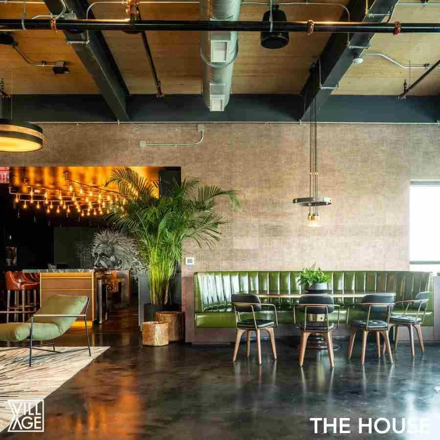 Interior of a modern restaurant with a green booth, a round table with a plant, and several chairs. There is a large potted plant and wall art of a lion in the background. Exposed ceiling pipes and ducts, with warm lighting and a window on the right.