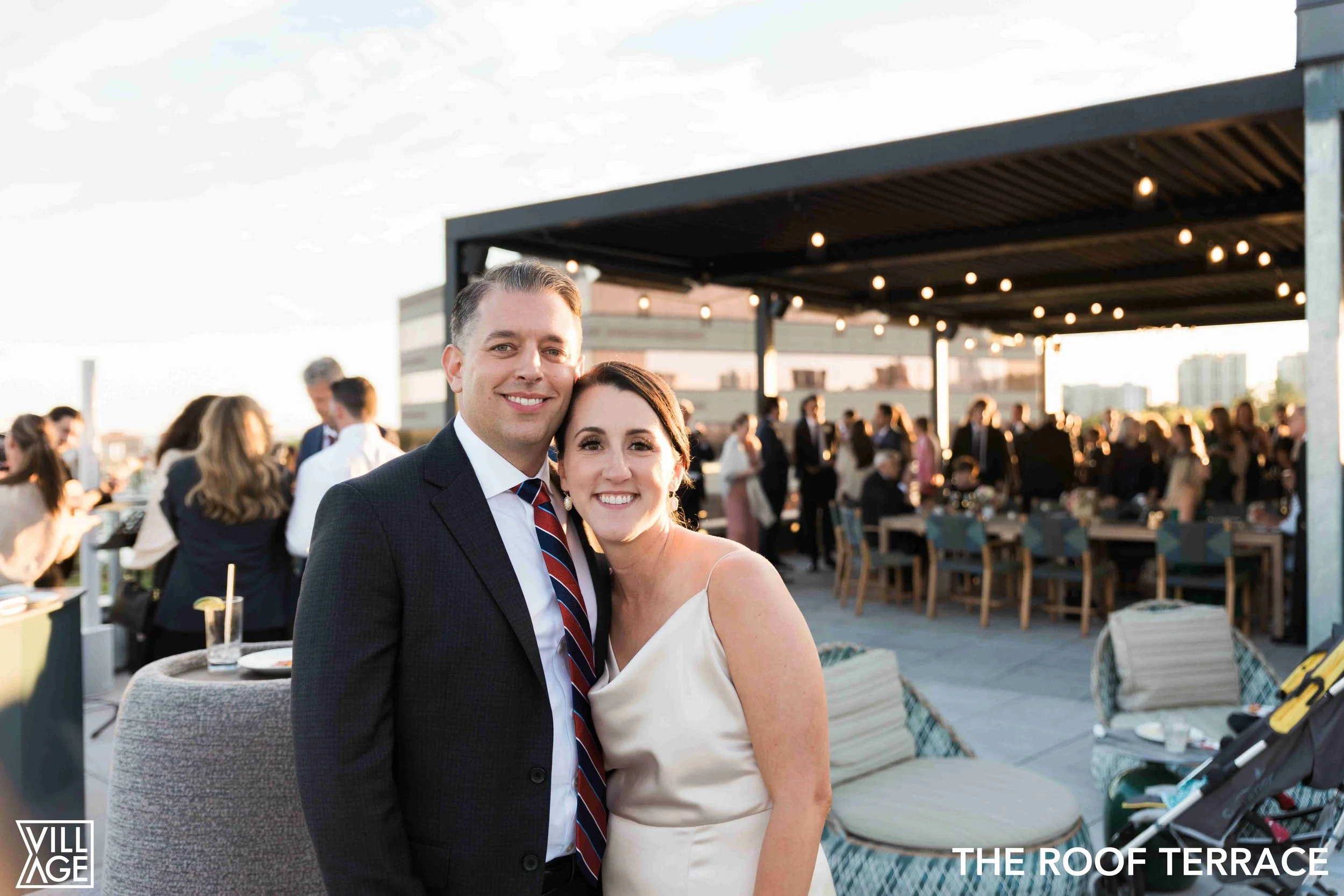 A smiling man and woman dressed in formal attire on a rooftop terrace during a social event at sunset, with string lights and many guests in the background.