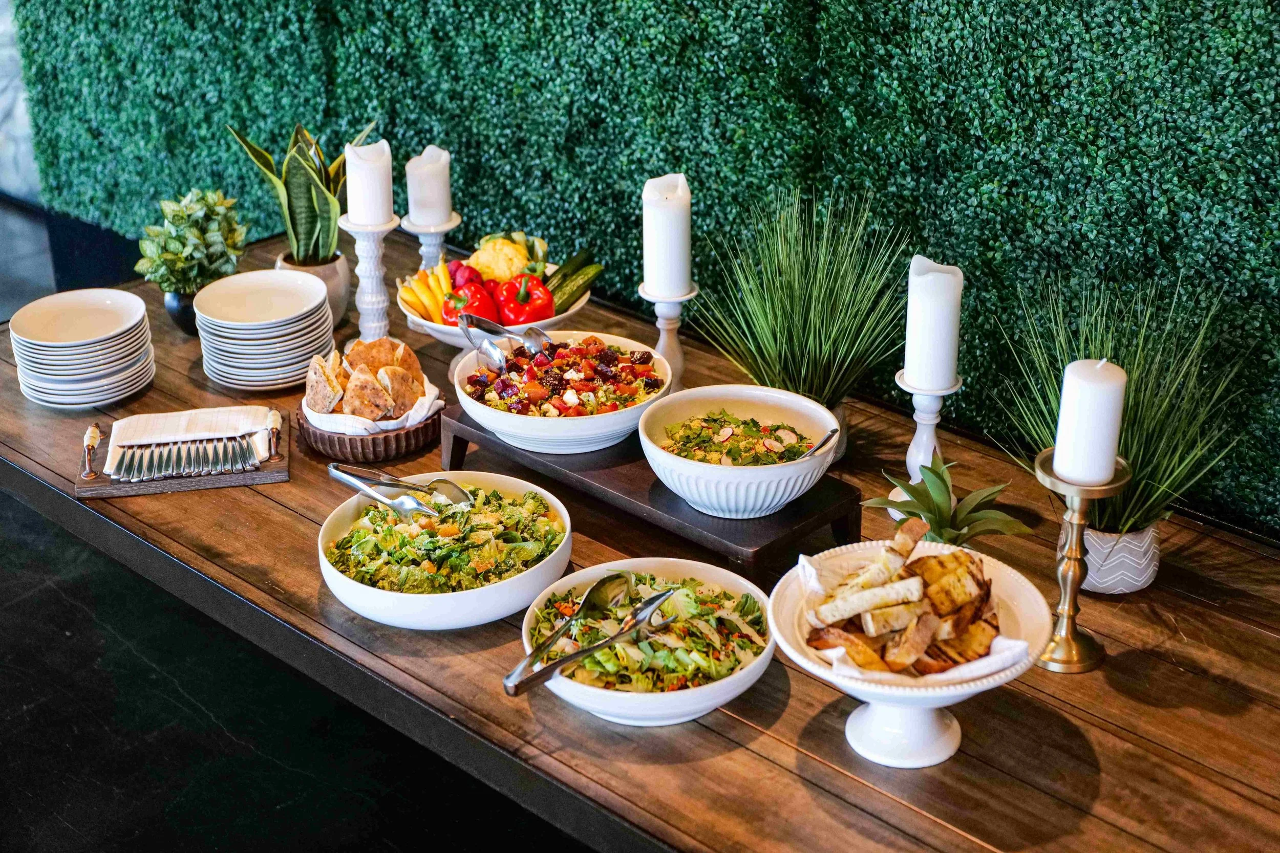 Assorted salads, bread, and vegetables on a wooden table with candles and decorative plants