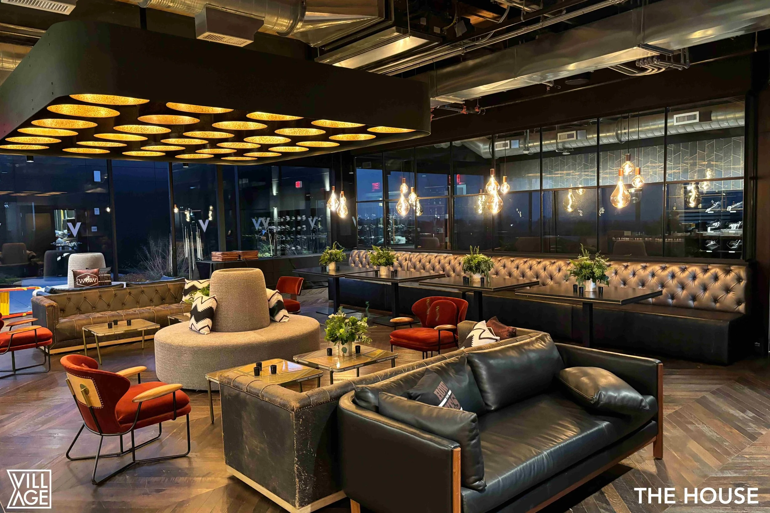 Interior of a modern lounge or restaurant featuring black leather sofa, red and beige armchairs, wood flooring, pendant lights, and plants, with floor-to-ceiling windows and creative ceiling lighting.