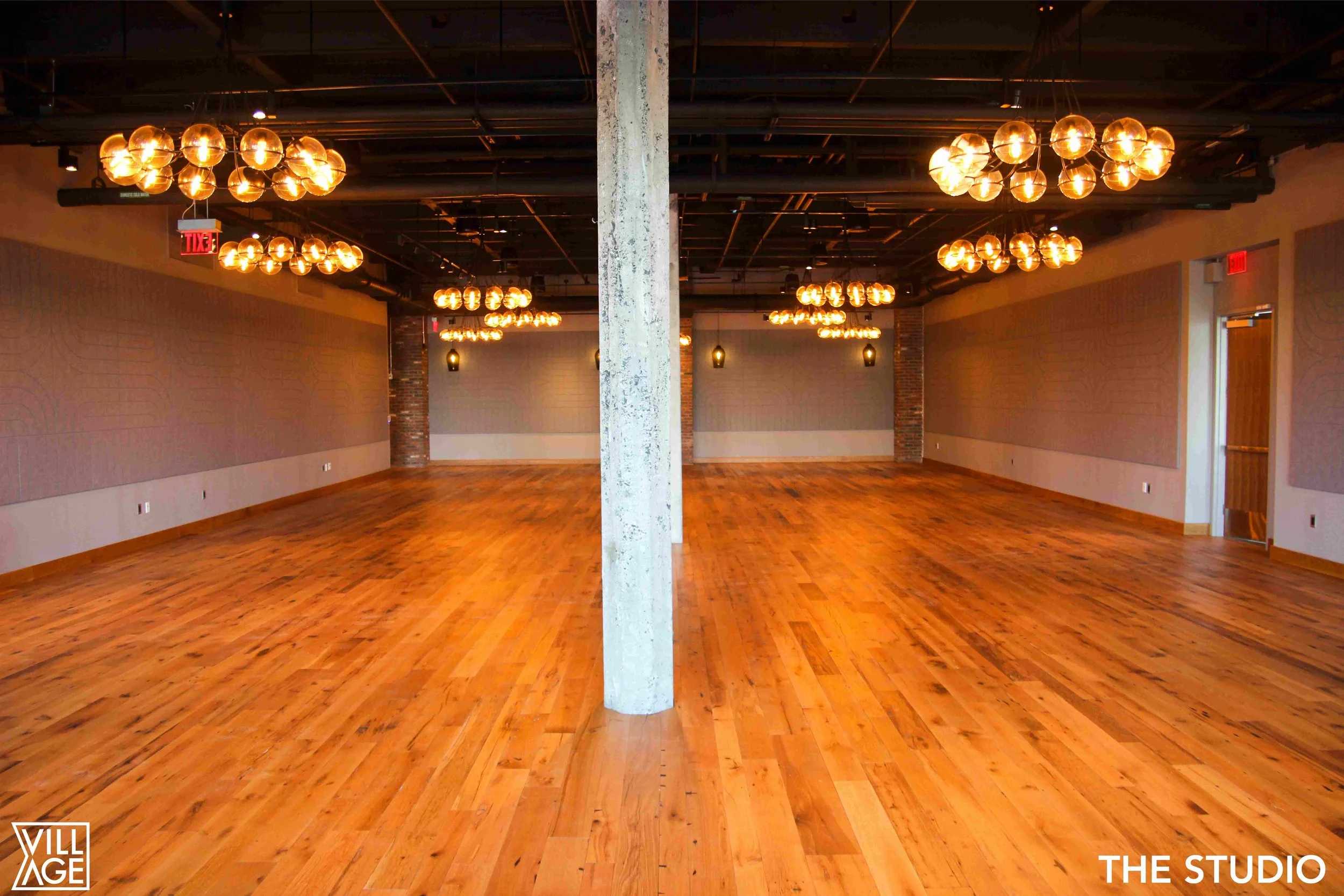 Empty event studio with wooden floors, exposed ceiling with black beams, and warm hanging light fixtures. There are brick columns, side wall panels, and a door on the right. The studio is well-lit and spacious.