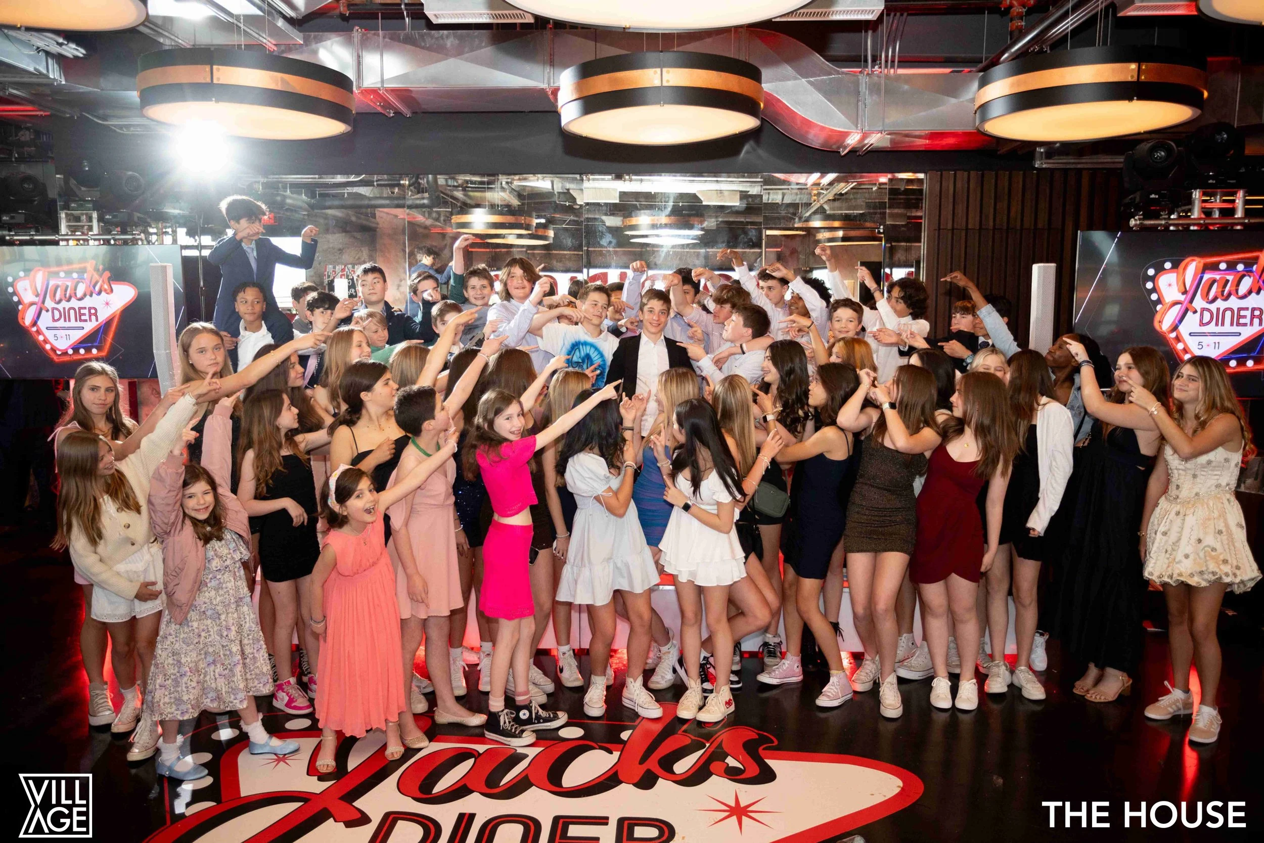 A large group of children and teenagers dancing and having fun in a dance studio or party room with a retro sign that reads 'Jack's Diner'. The studio features a mirrored wall, circular ceiling lights, and a logo in the lower left corner that says 'V