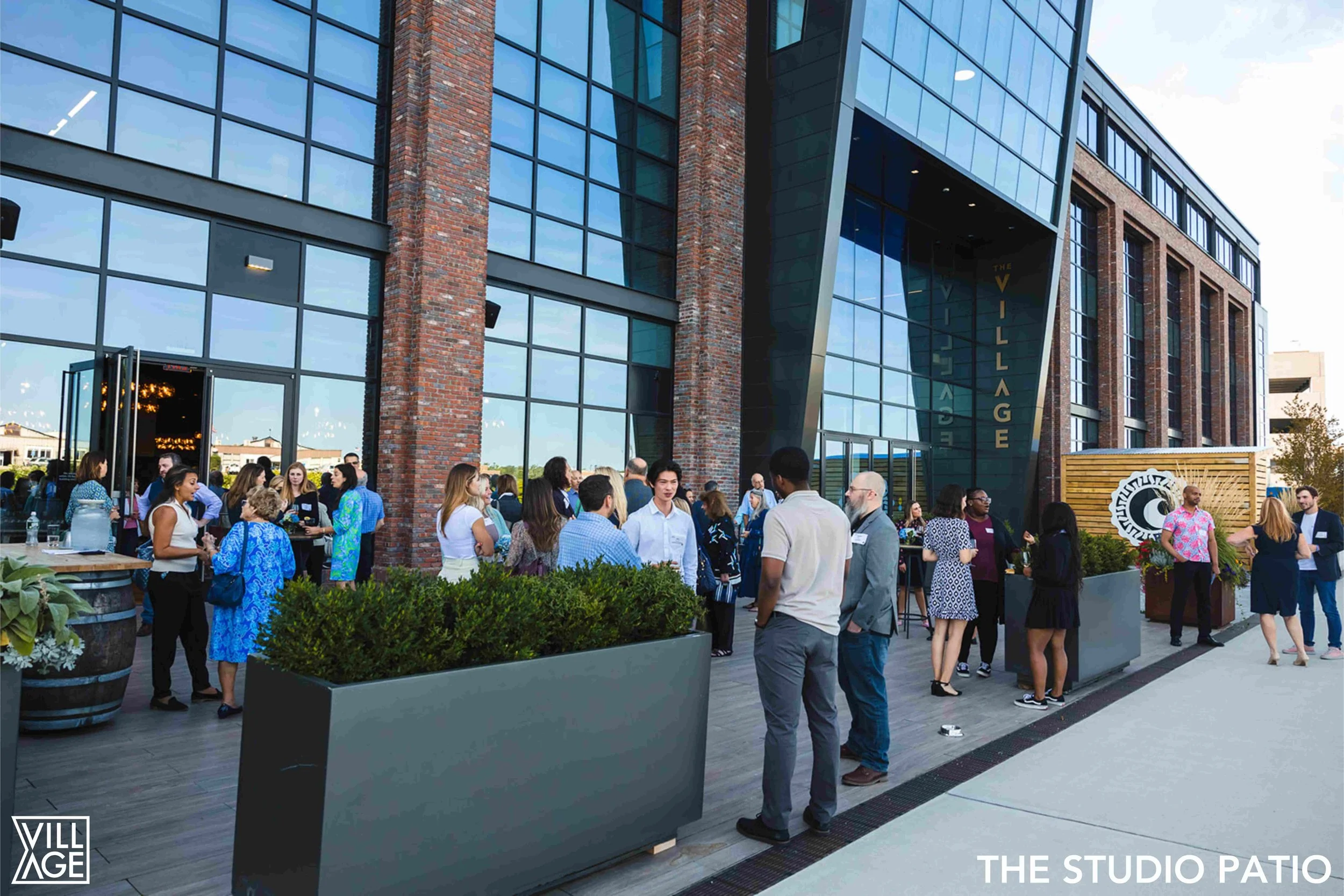 People gathering outside a modern building labeled 'The Village' at the studio patio, with large glass windows, brick accents, and landscaped planters, during a social event.