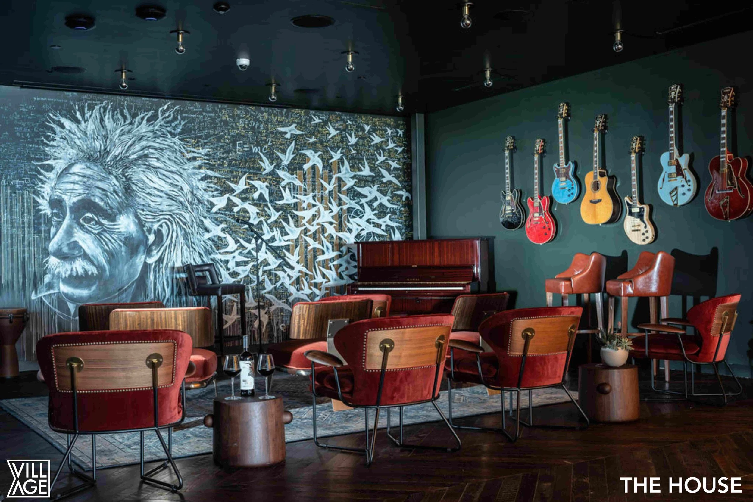 A music room with a mural of Albert Einstein's face and a flock of birds on the wall, a row of colorful guitars hanging, and chairs arranged around a piano in the center.