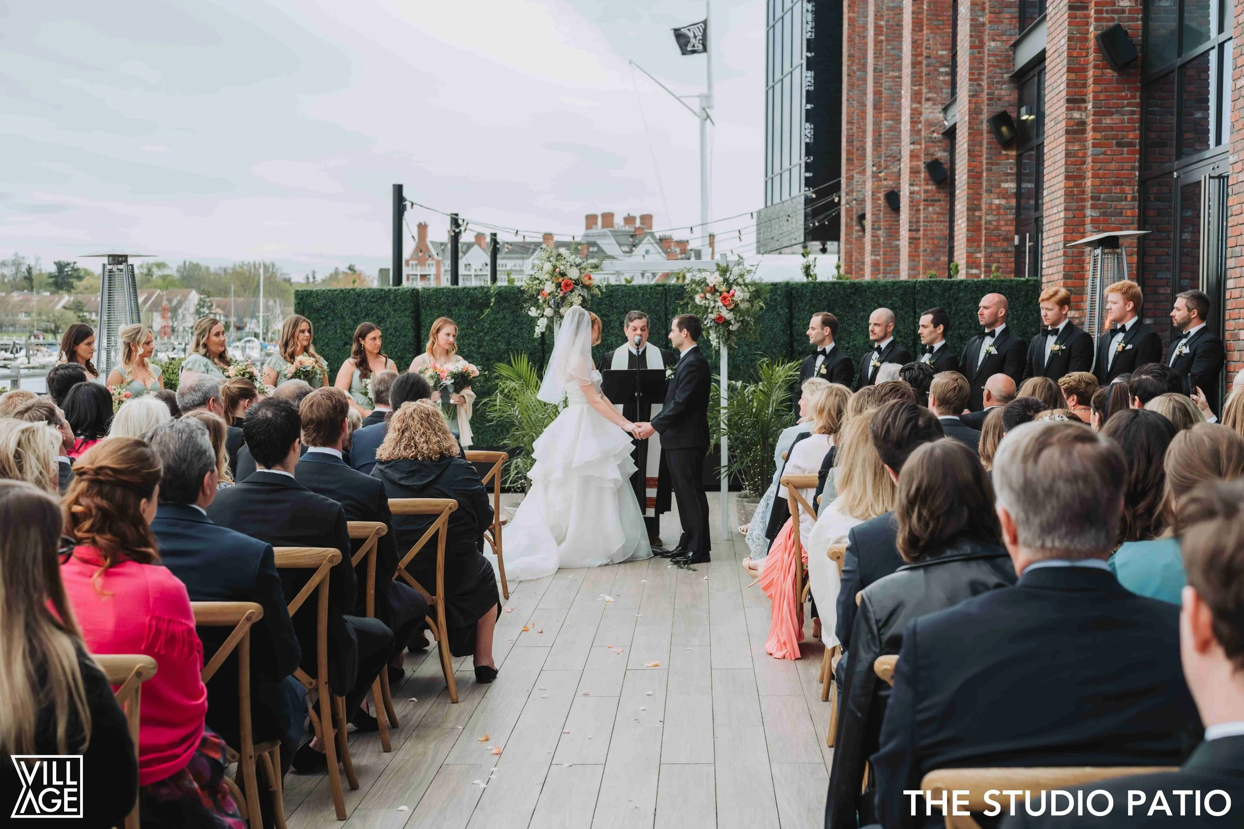 Outdoor wedding ceremony on a patio with a bride and groom exchanging vows, surrounded by bridesmaids and groomsmen, with seated guests and cityscape in the background.