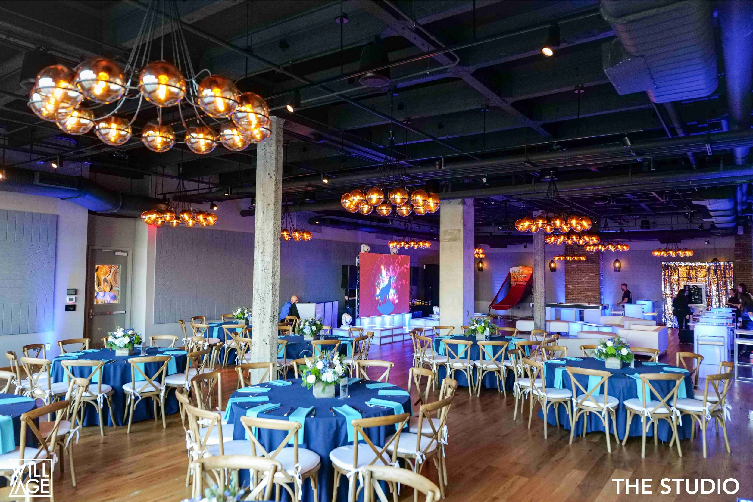 Event space with round tables covered in blue tablecloths, decorated with white and blue flower centerpieces, set with blue napkins, surrounded by wooden chairs. The room has hanging amber-colored light fixtures, wooden flooring, concrete columns, an