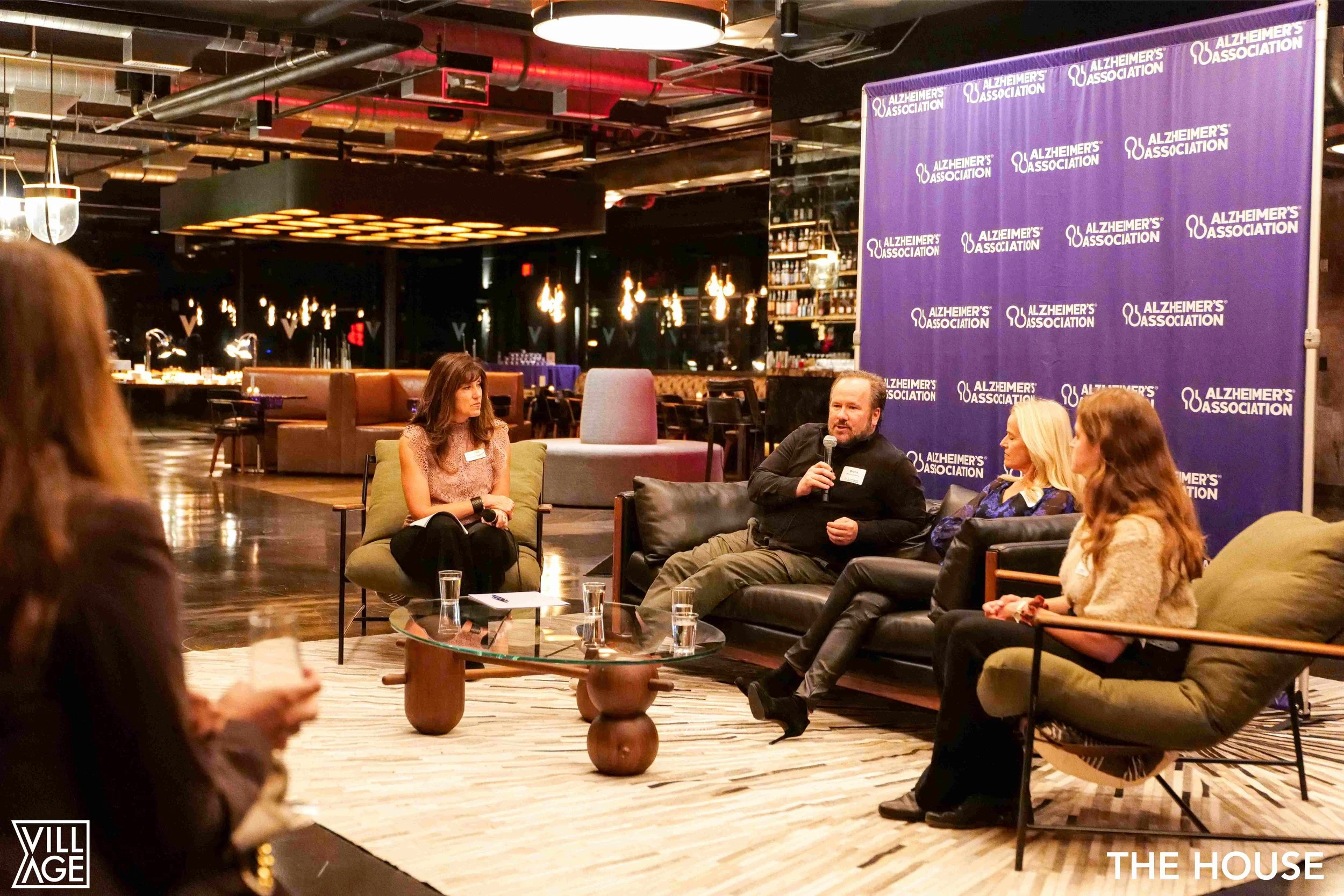 Panel discussion at an Alzheimer's Association event in a modern, dimly lit venue with four women and one man seated on sofas and chairs, a purple backdrop with the Alzheimer's Association logo, and a bar area in the background.