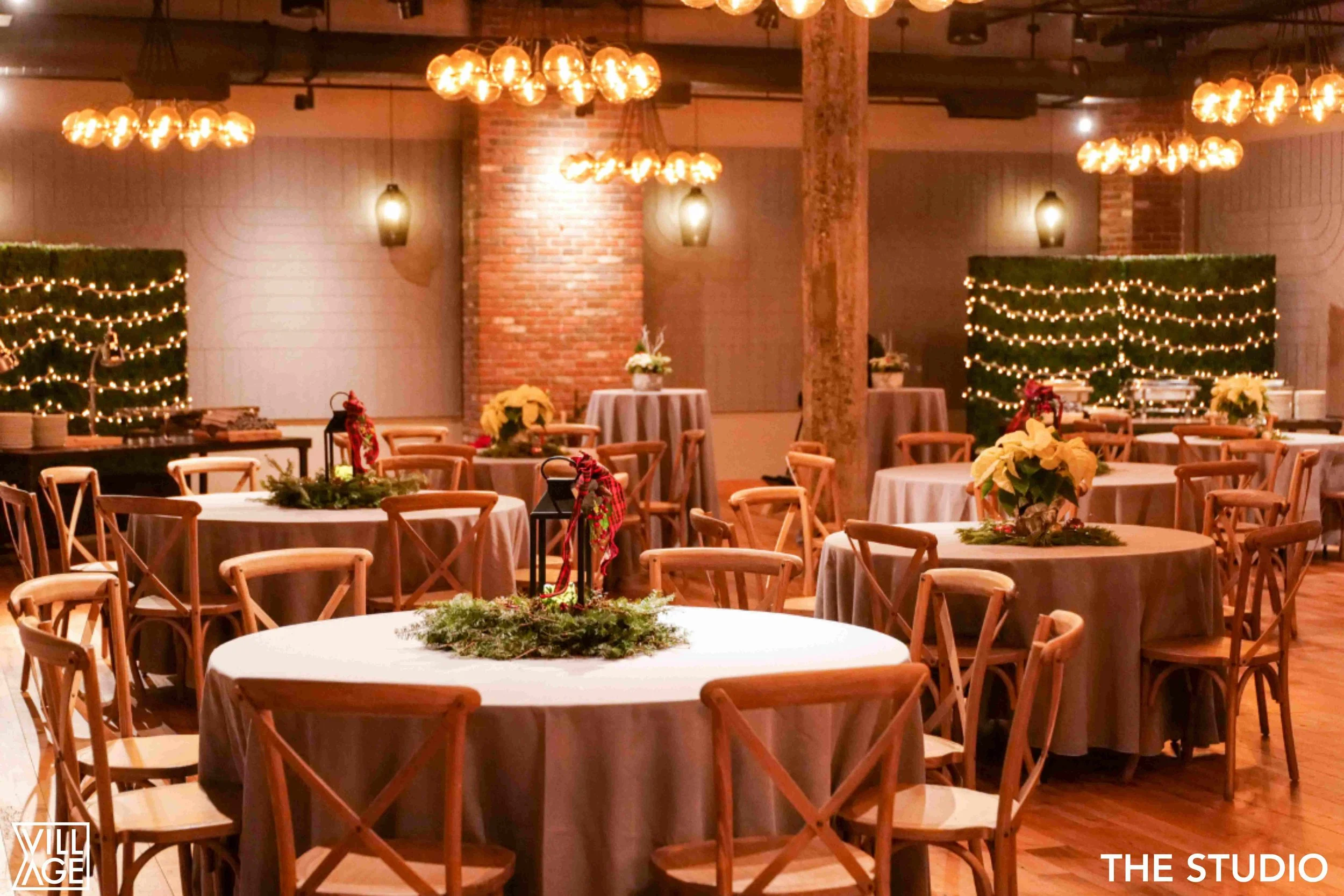 A festively decorated event space with round tables covered in dark tablecloths, adorned with Christmas centerpieces featuring poinsettias and greenery. Wooden chairs surround the tables. The background includes warm lighting fixtures, brick walls, a