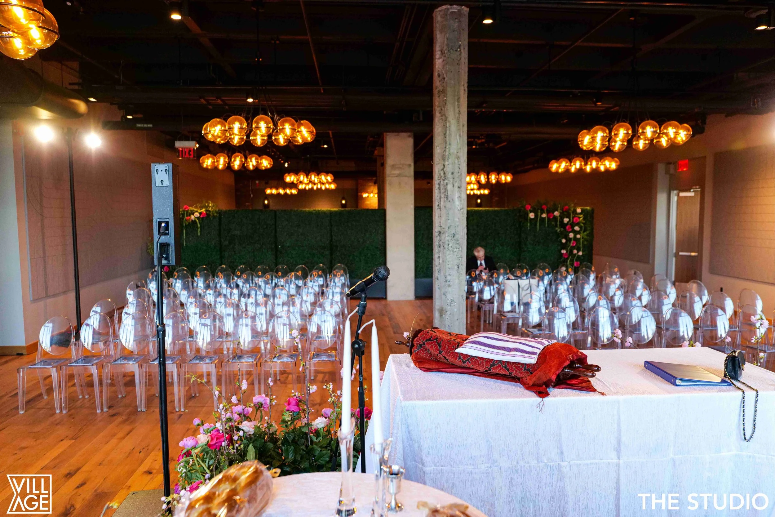 Wedding or event space with rows of clear acrylic chairs, floral decorations, and a panel of green hedge with pink flowers. A table with a rolled fabric, book, and purse is in the foreground. The room features warm lighting and a microphone setup nea