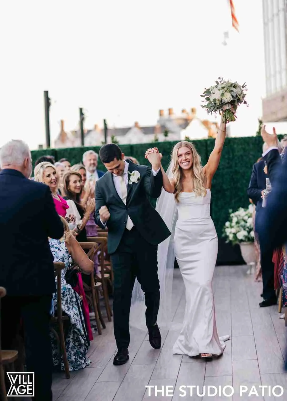 A joyful bride and groom celebrating with guests at their wedding reception. The bride holds a bouquet in one hand and raises her hand in victory, while the groom smiles and looks down, walking alongside her. Guests are clapping and cheering around t