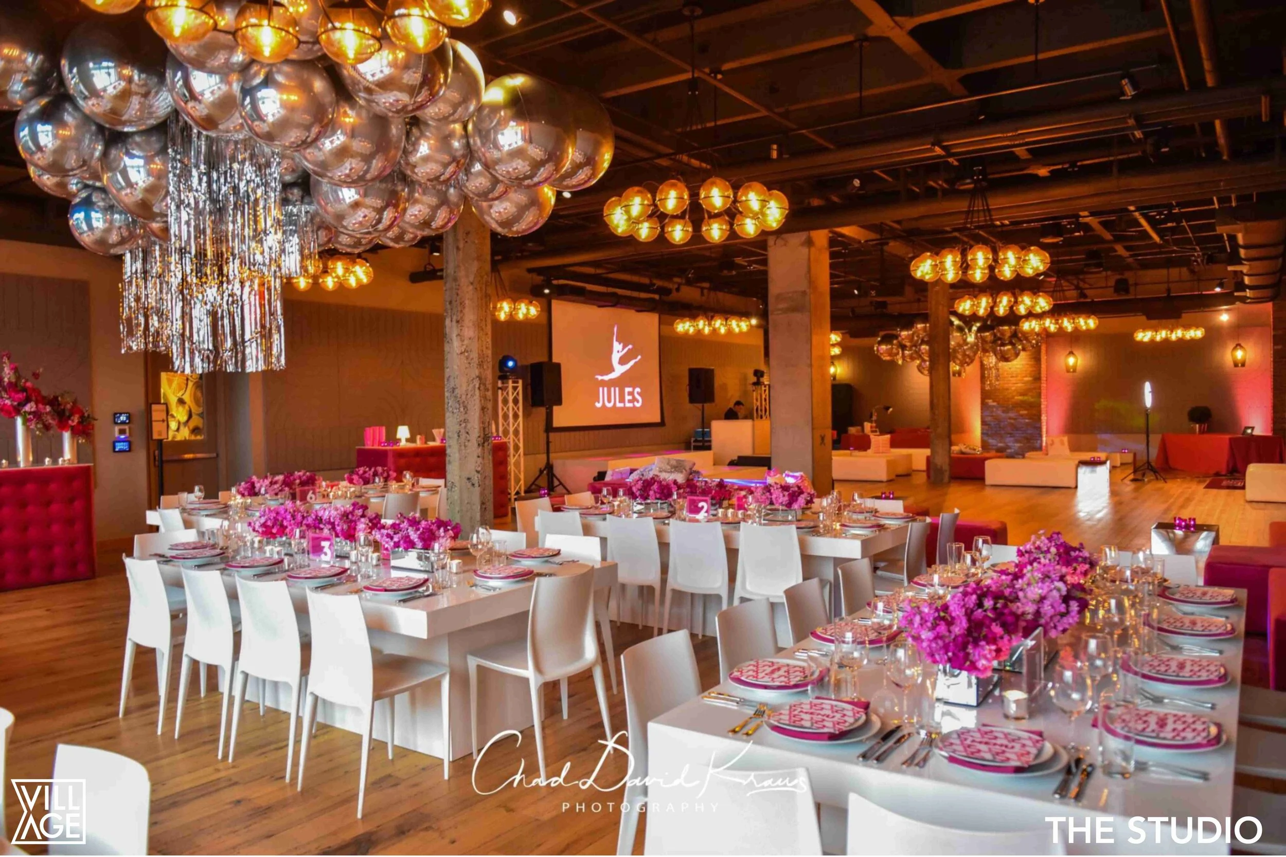 Upscale event space decorated with pink flowers on tables, hanging metallic balloons, and warm ambient lighting, ready for a celebration.