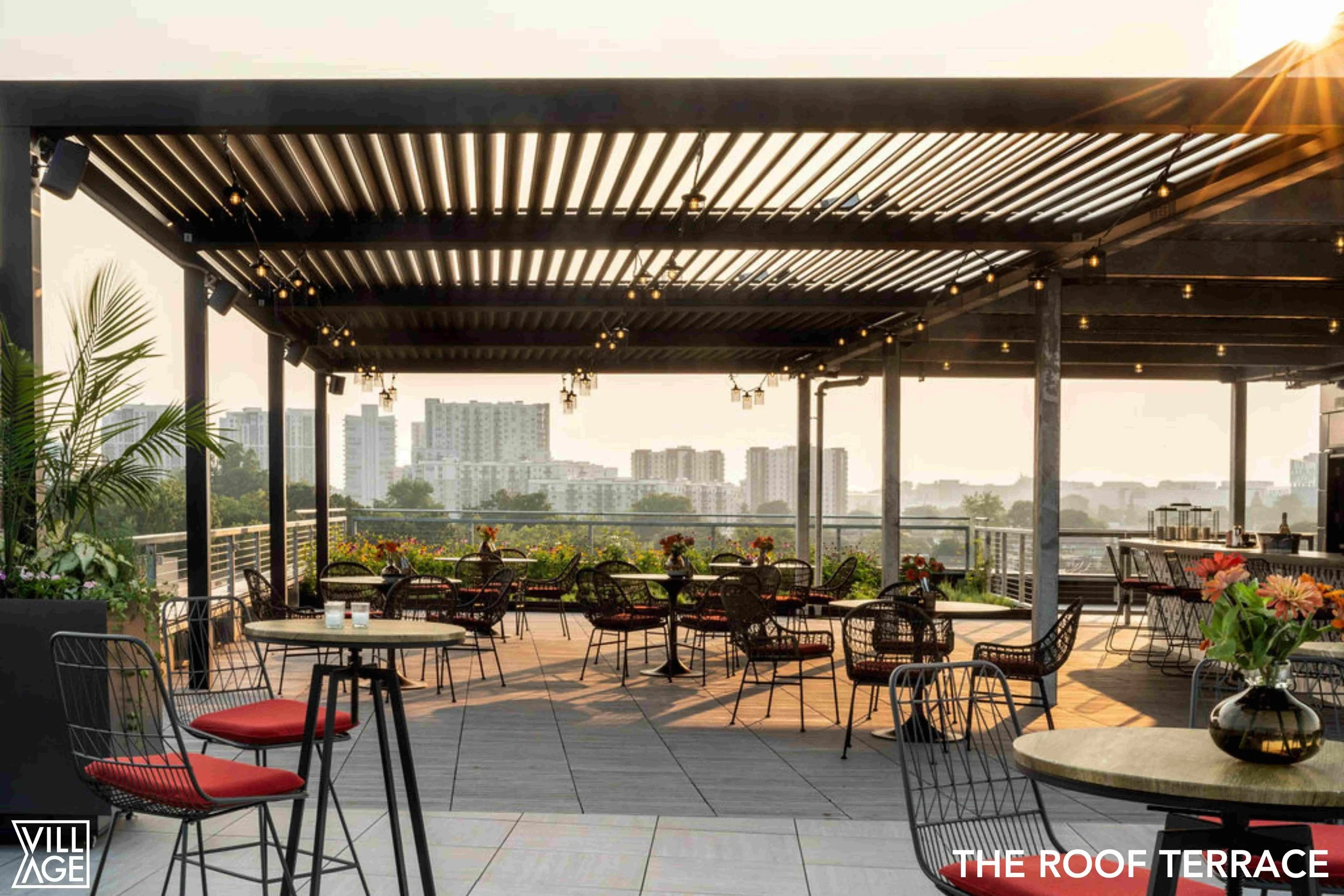 An outdoor roof terrace with tables and chairs, decorated with flowers, overlooking a city skyline at sunset.