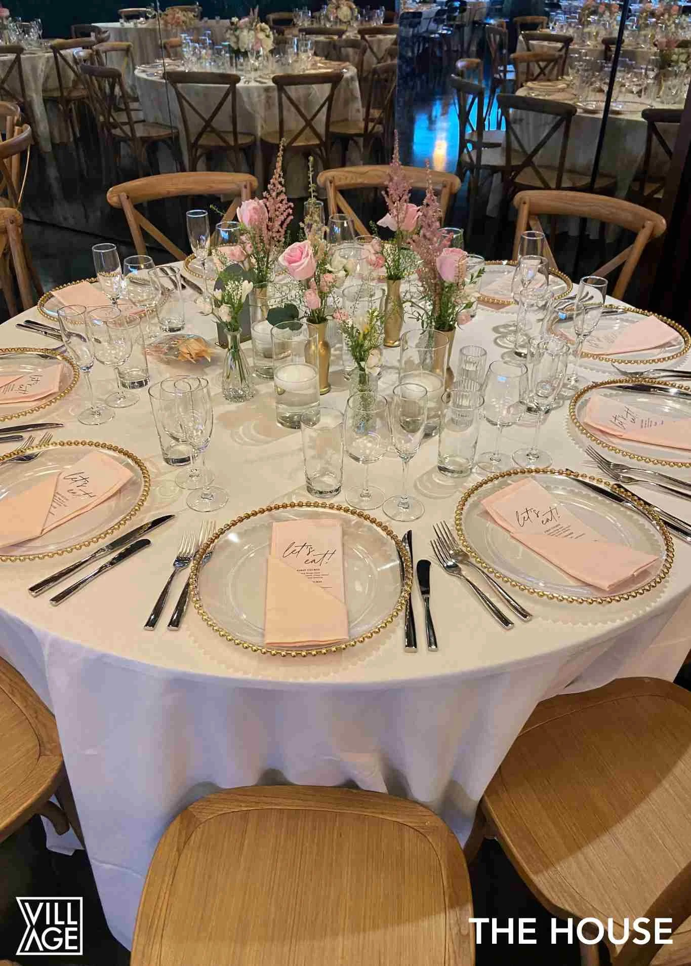 A round banquet table decorated for a formal event, with pink and white floral centerpieces and place settings including gold-trimmed clear plates with pink napkins and handwritten messages, surrounded by wooden chairs and multiple glasses.