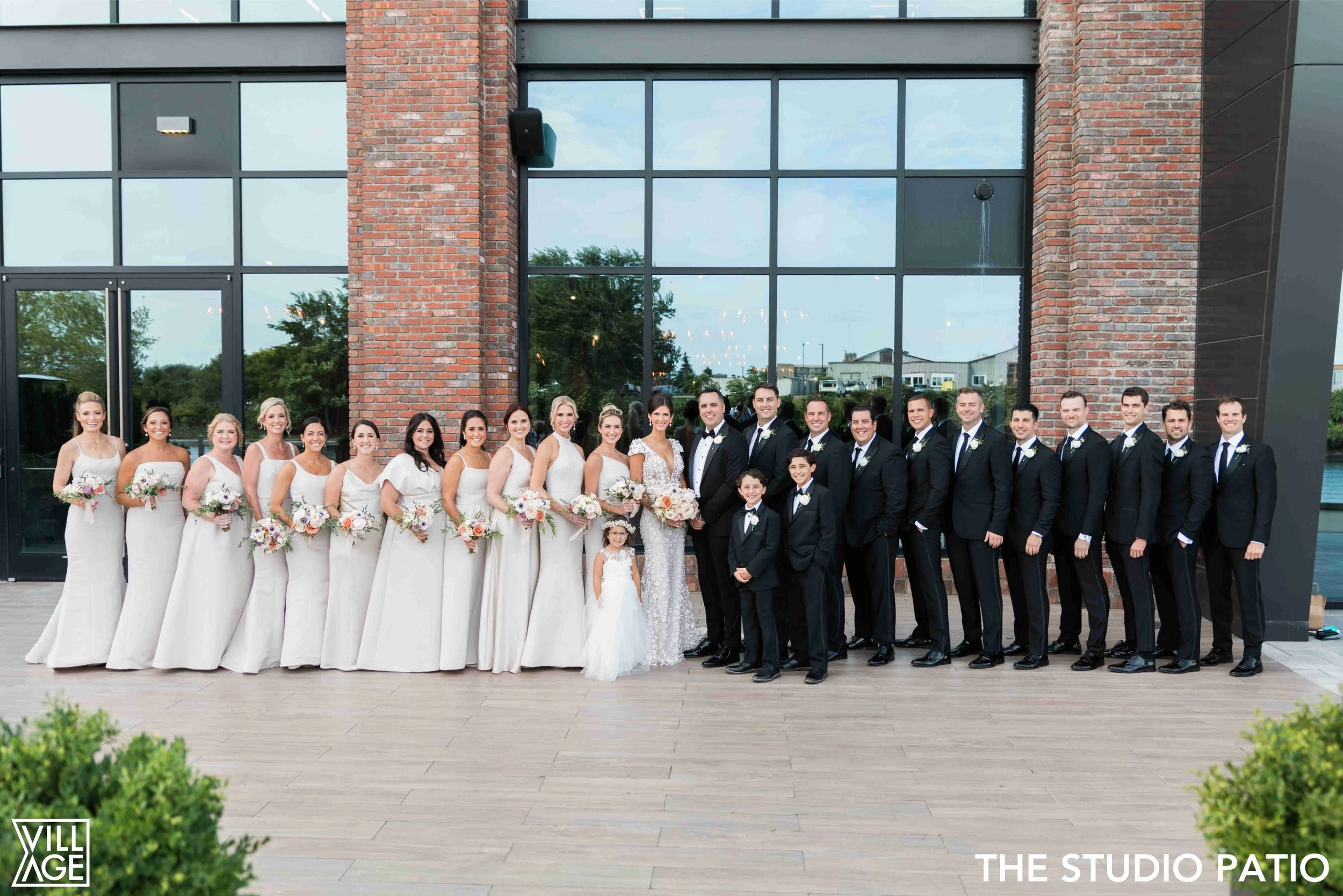 Group of wedding party members dressed in formal attire standing outside in front of a large glass window and brick wall. The group includes bridesmaids in cream dresses holding bouquets, groomsmen in black tuxedos, a bride in a white wedding dress, 