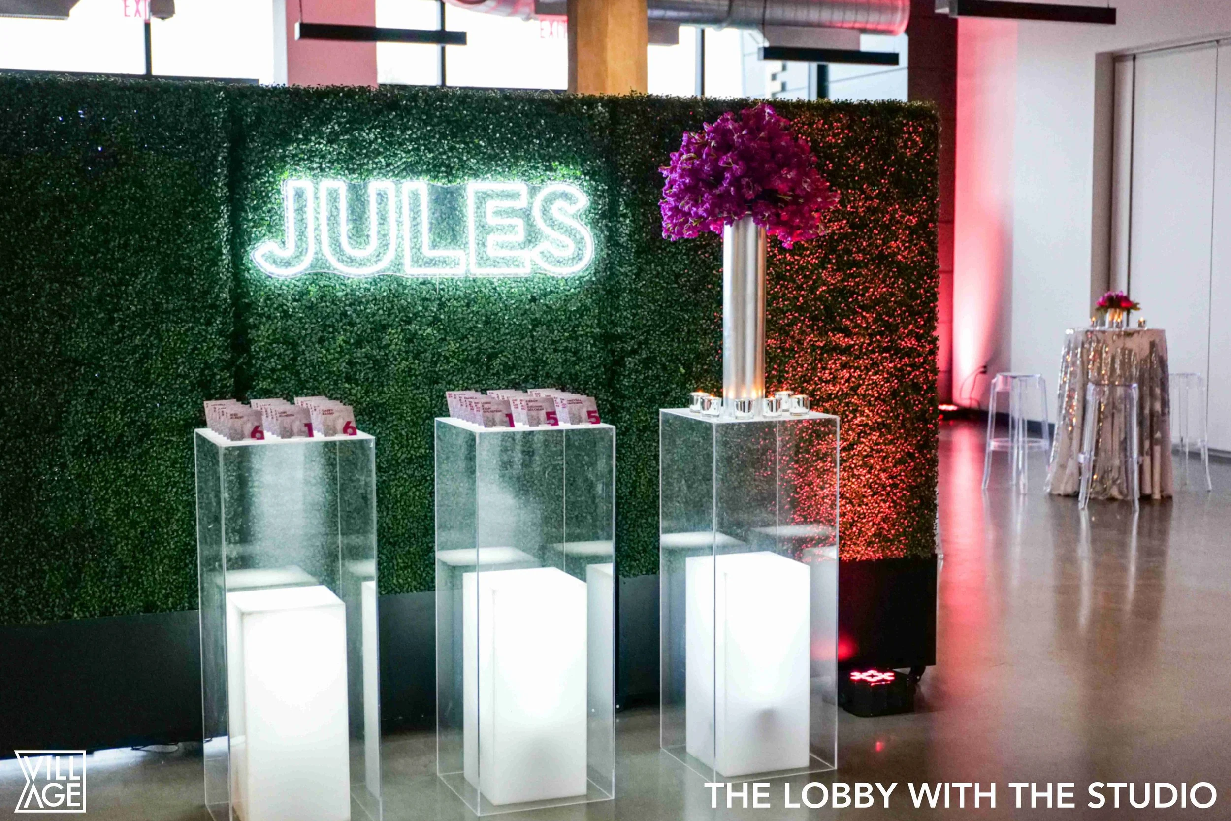 Event lobby with a green hedge wall backdrop, illuminated neon sign reading 'JULES,' clear display stands with pink event tickets, a tall floral arrangement with pink flowers, and a nearby table with a floral centerpiece. The space has modern decor a