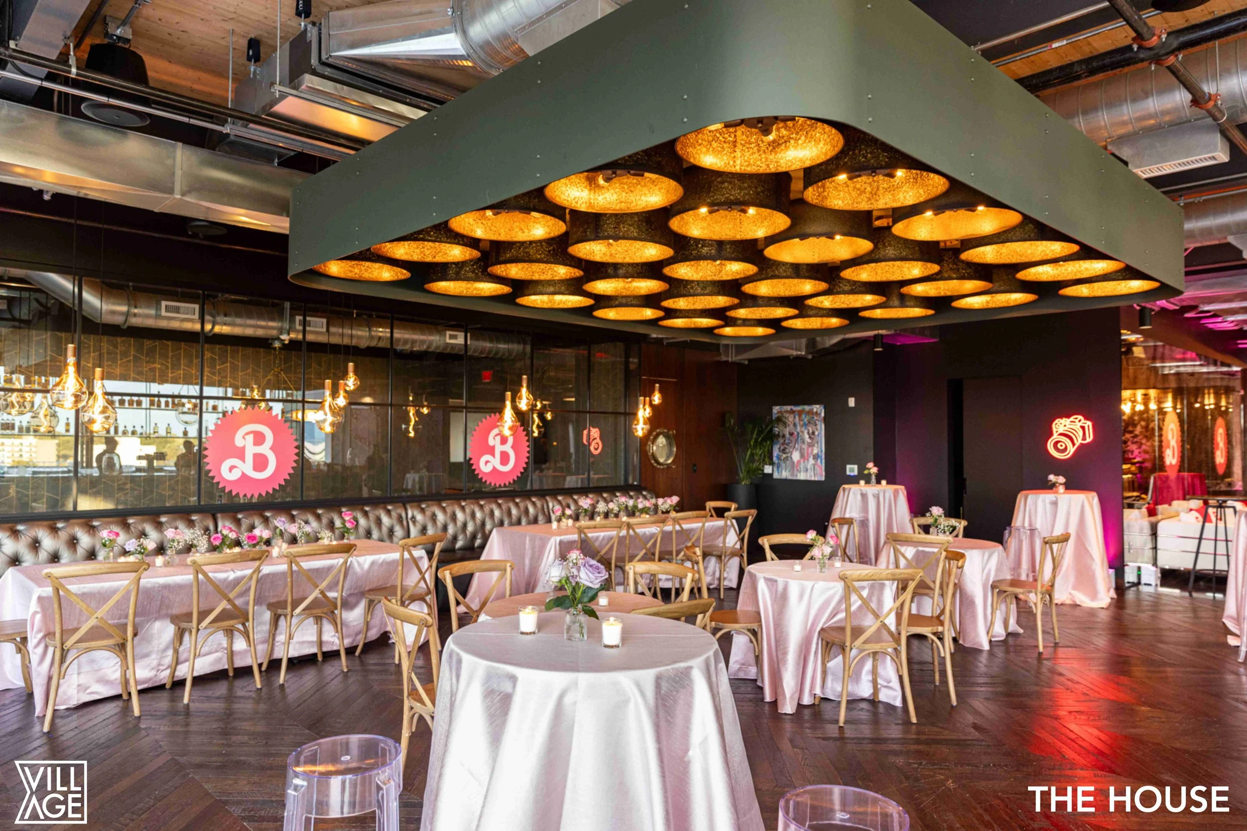 Upscale restaurant interior with pink tablecloths, wooden chairs, and decorative lighting, featuring a large ceiling fixture with round gold lights and pink neon signs.