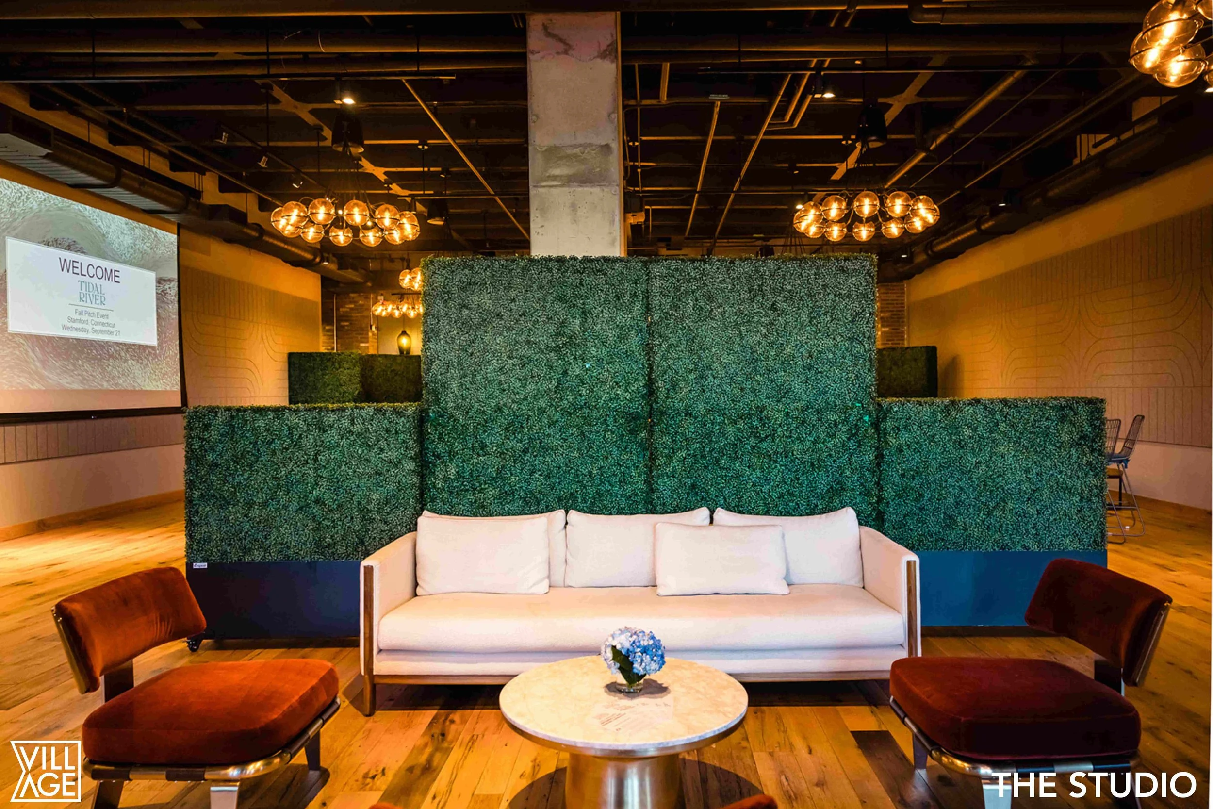 Interior of a modern lounge with a white sofa, two brown chairs, a small round table with a flower arrangement, a large green artificial hedge wall, hanging spherical lights, and a large screen displaying a welcome message in the background.