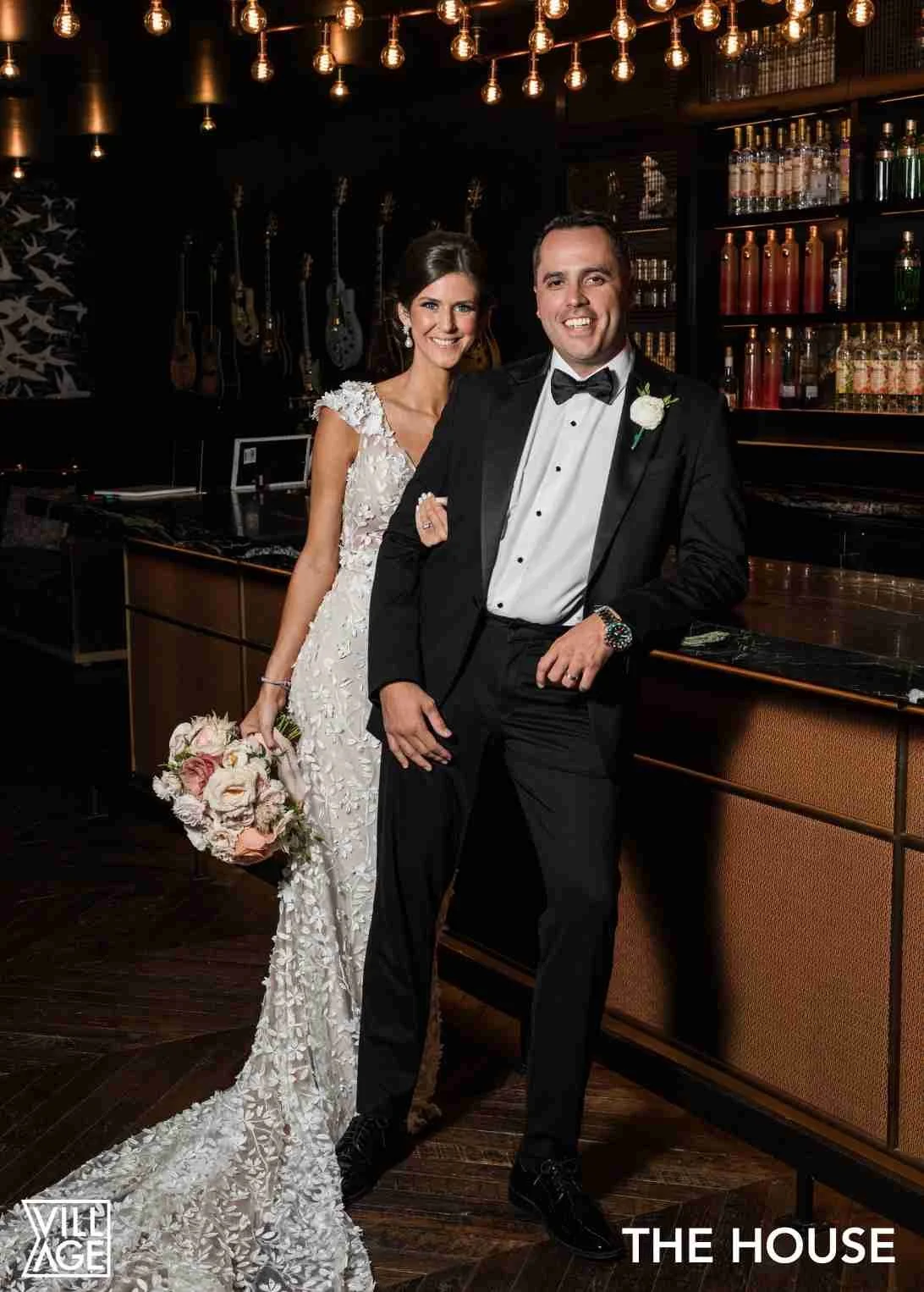 A bride and groom in formal wedding attire inside a stylish bar or lounge with guitars hanging on the wall and warm hanging light bulbs.