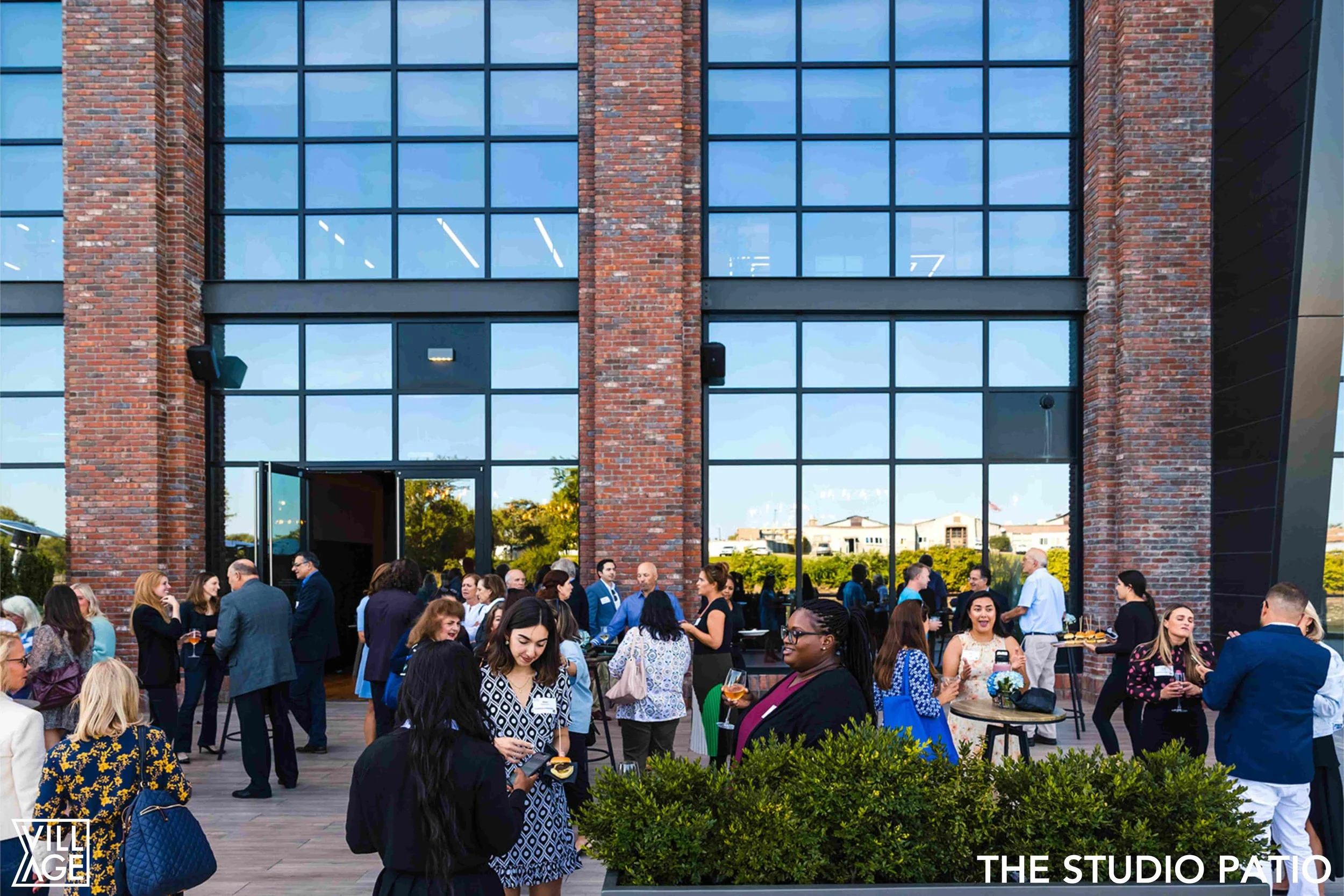 People socializing at an outdoor patio event in front of a modern brick building with large glass windows.
