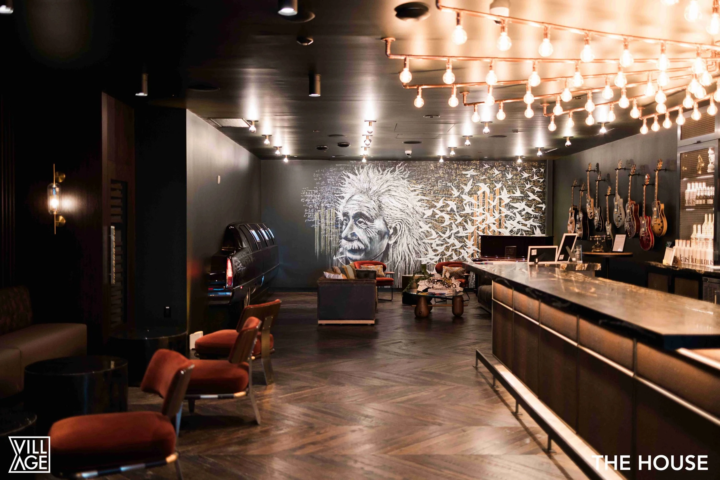 Interior of a modern bar or lounge with black walls, a large mural of Albert Einstein, a lit chandelier, and guitars hanging on the wall.