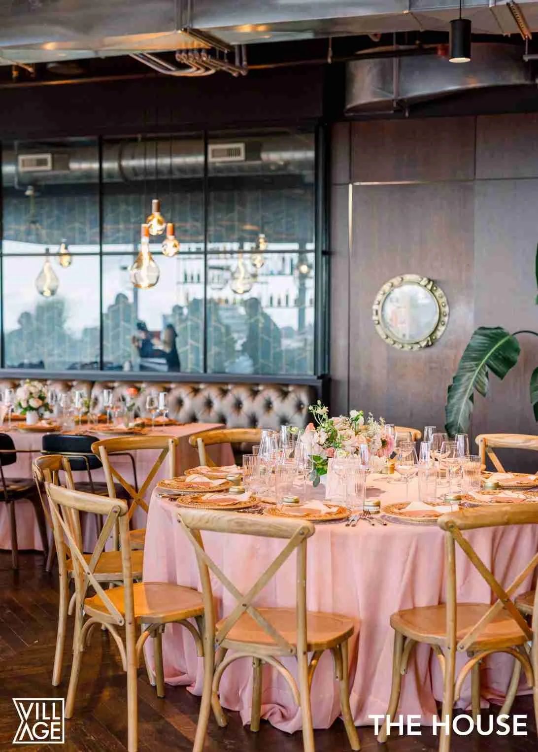Elegant banquet table set with pink tablecloth, floral centerpieces, glassware, and place settings, in a modern restaurant with large windows, hanging light bulbs, dark walls, and lush green plants.