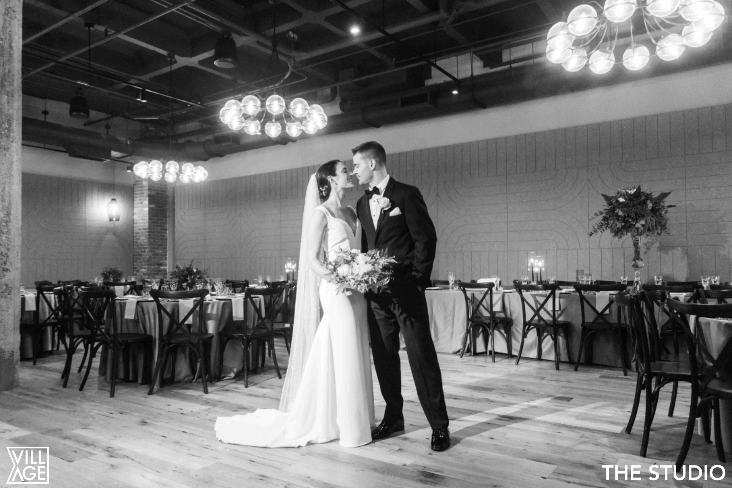 Black and white photo of a bride and groom standing close together in a decorated wedding reception hall, with tables and chairs in the background, and large floral arrangements on the tables.
