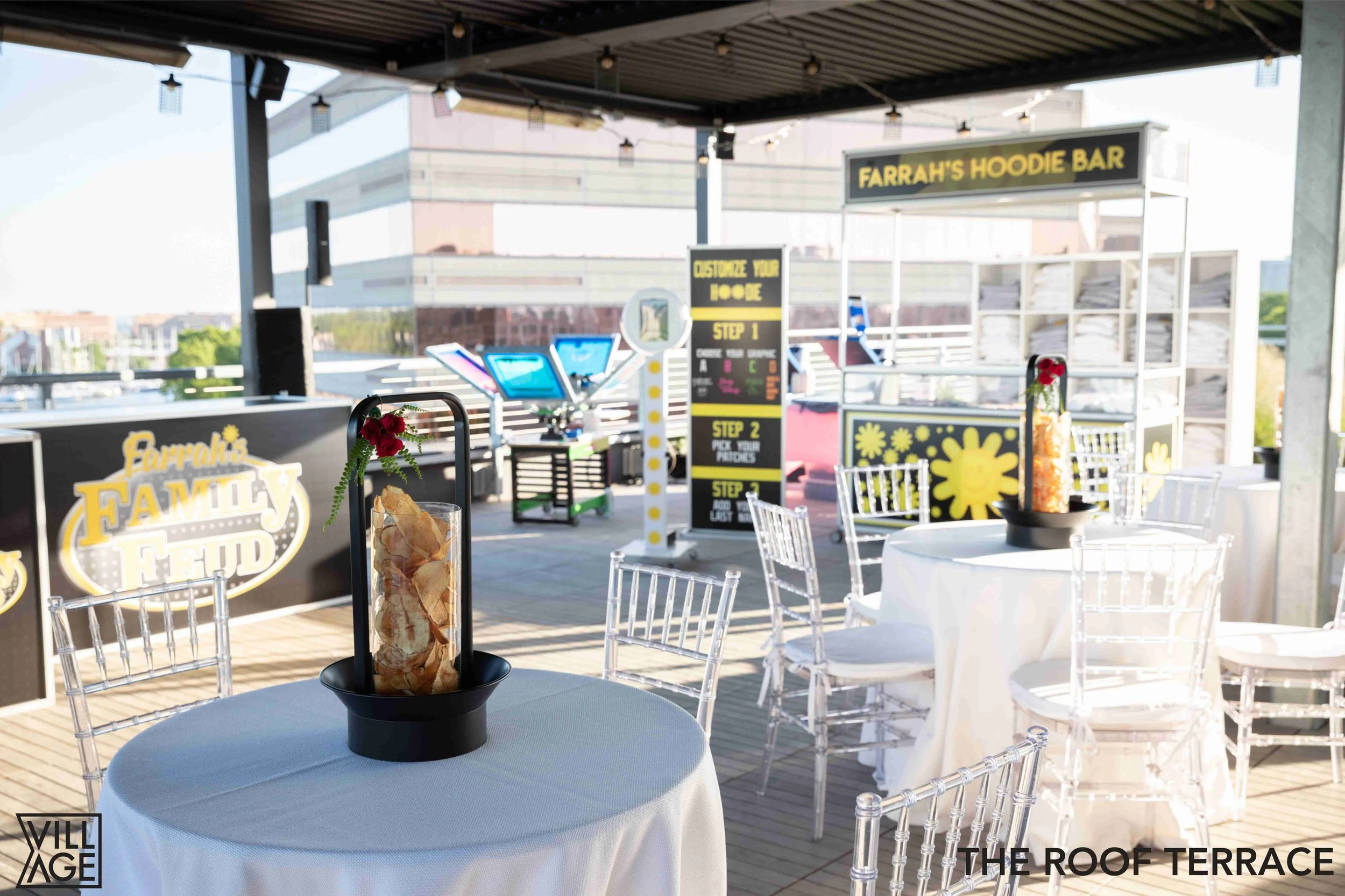 Outdoor roof terrace with round tables covered in white tablecloths, transparent chairs, floral centerpieces, a food stand labeled 'Farrah's Hood Bar', and a chalkboard menu with steps for ordering.