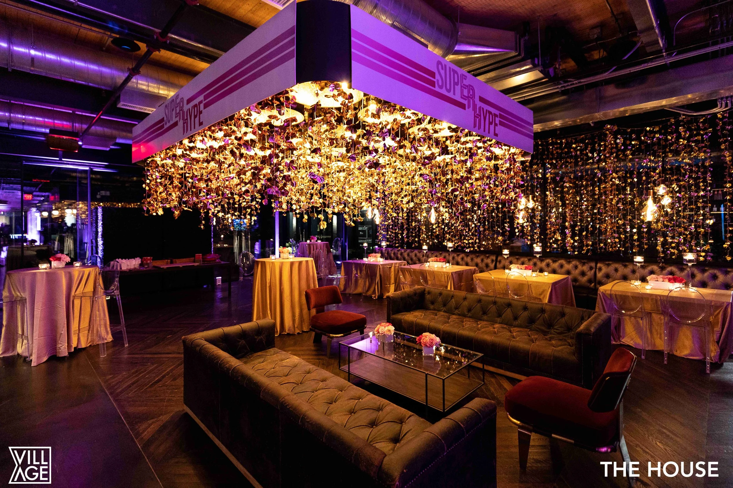 An event space decorated with purple and gold, featuring a hanging installation of gold leaves and lights under a sign that reads "Super Hype." There are sofas, tables with floral arrangements, and chairs, with purple and pink lighting creating a lux