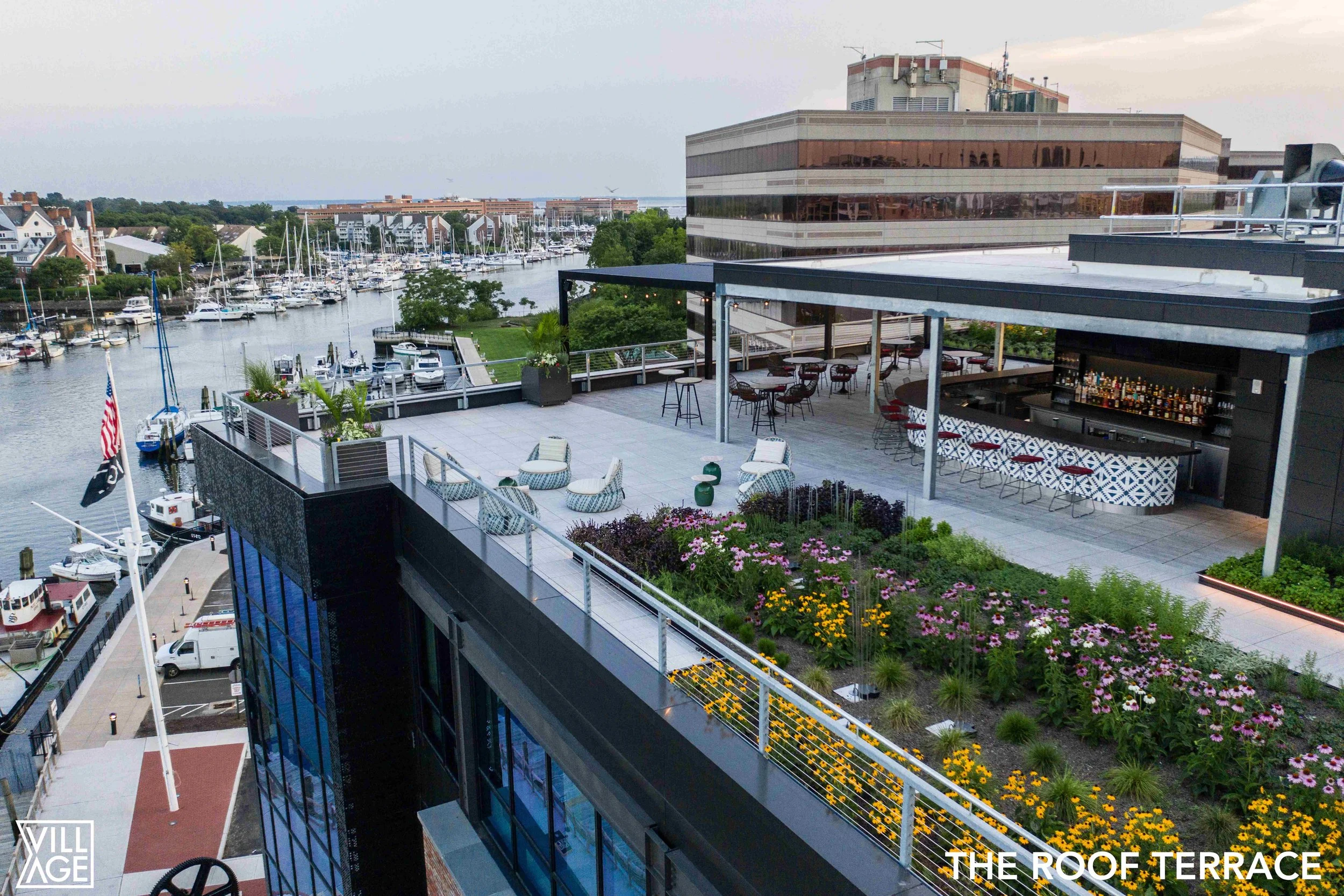 A rooftop terrace with outdoor seating, bar, and garden, overlooking a marina with sailboats and water, during daytime.