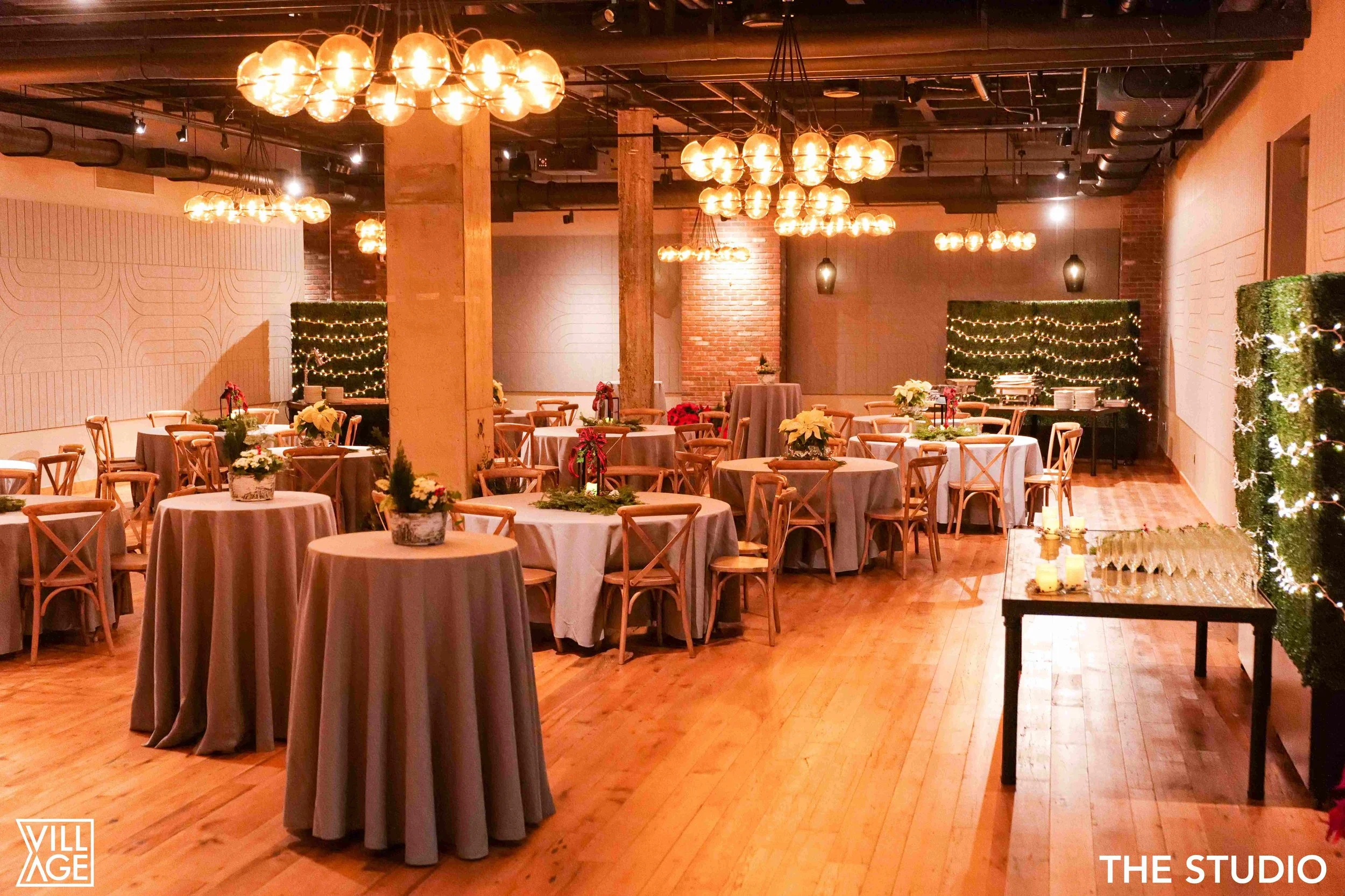 A decorated event space with round tables covered with gray tablecloths, wooden chairs, and holiday decorations, including poinsettias and string lights, set in a warm lighting ambiance with hanging globe lights and brick walls.